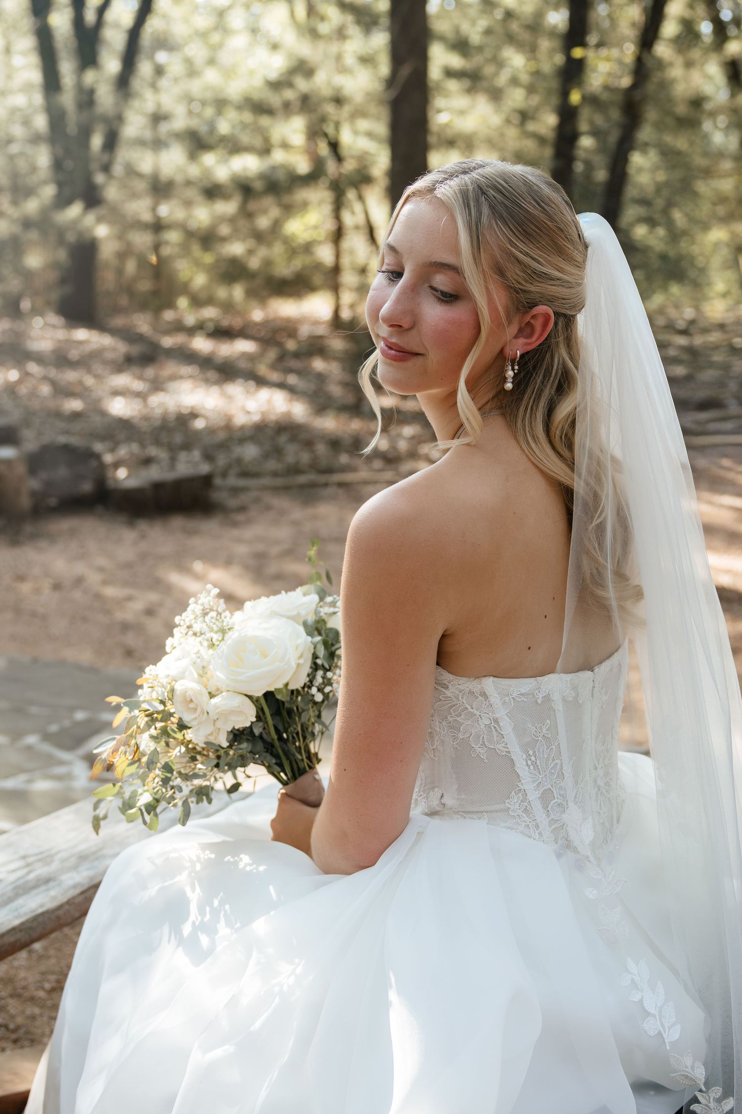 Elegant blonde bride in strapless lace wedding gown with veil holds white bouquet in wooded setting.