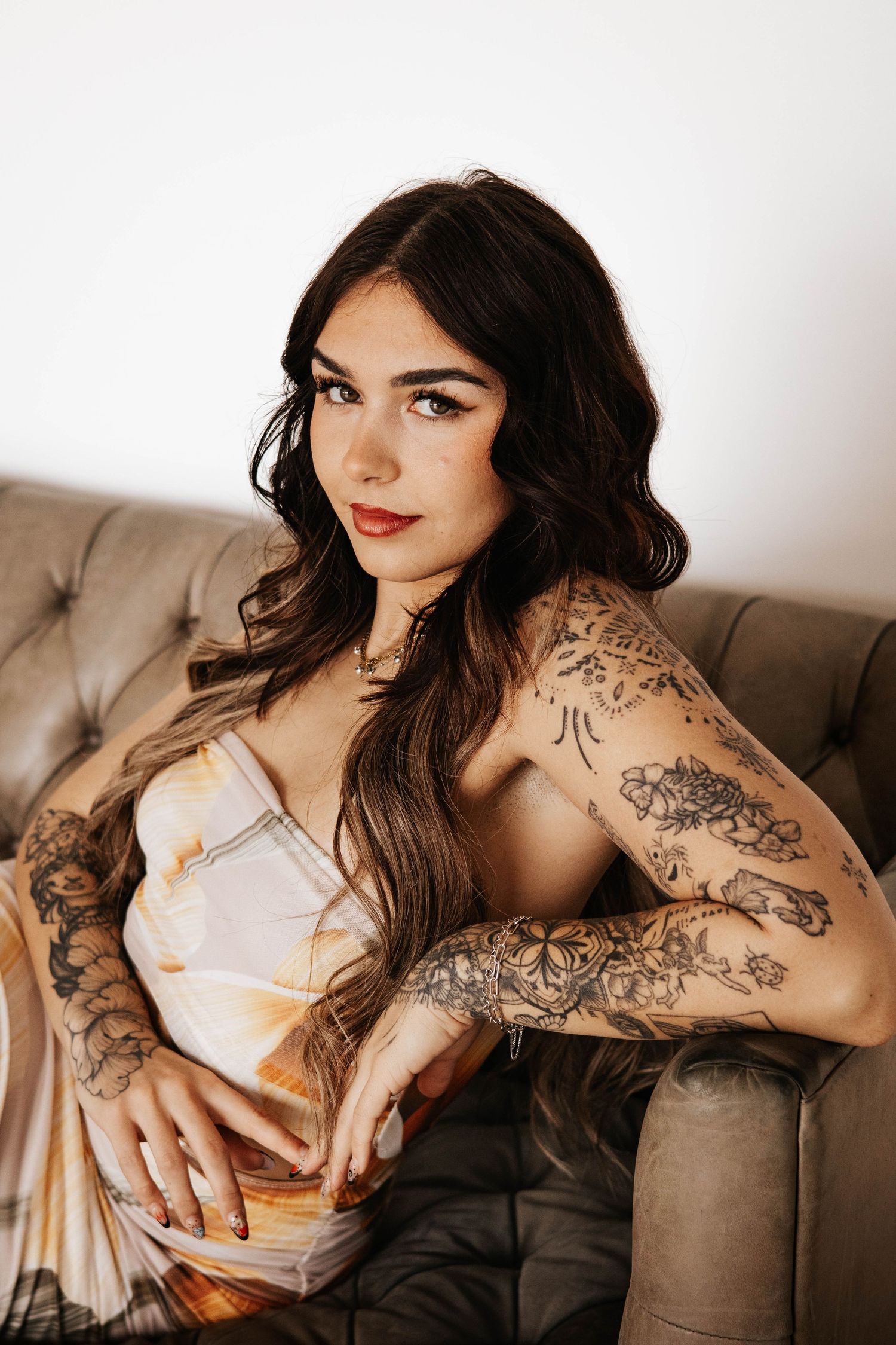 Woman in white dress with intricate arm tattoos poses on brown leather couch.