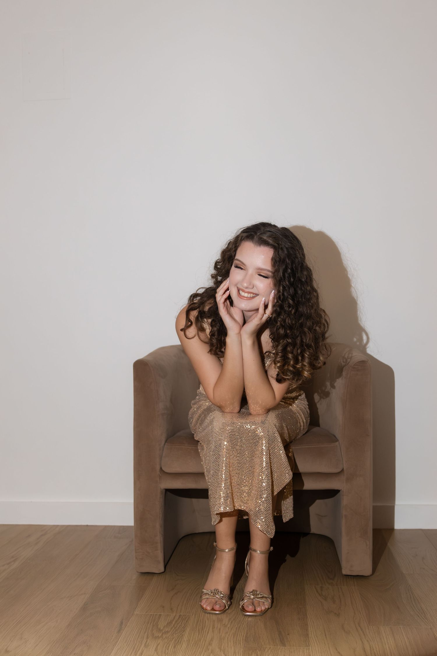 Smiling young woman with curly hair in a gold sequin dress sitting in a velvet chair.