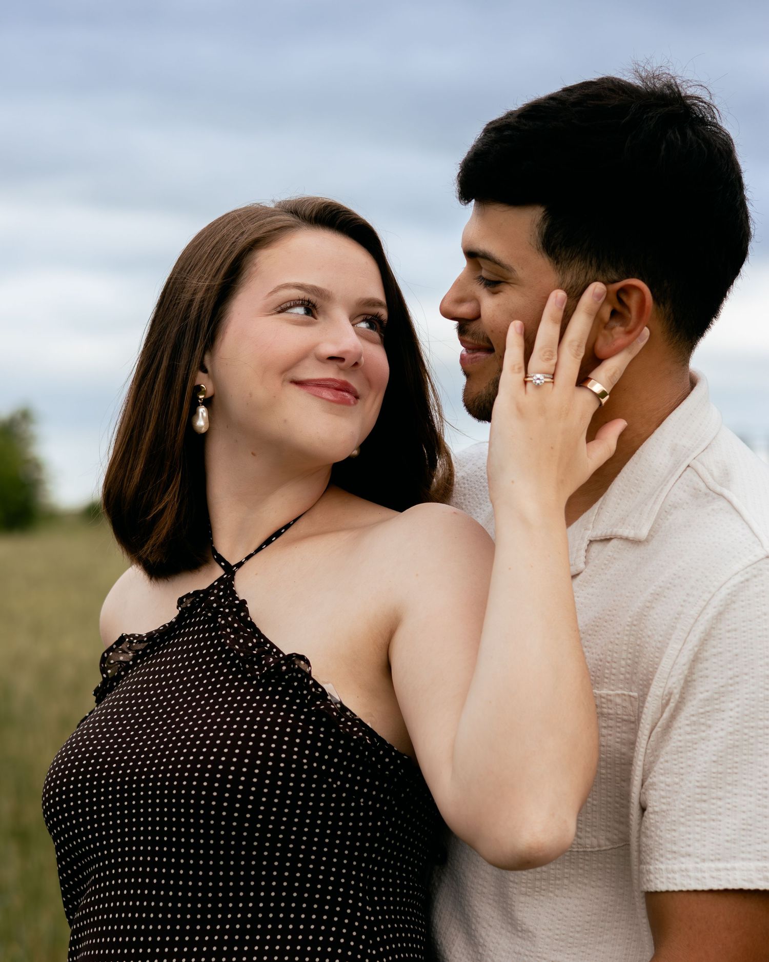 Couple sharing a romantic moment in a field, woman in black polka dot dress touching mans face with engagement ring visible.