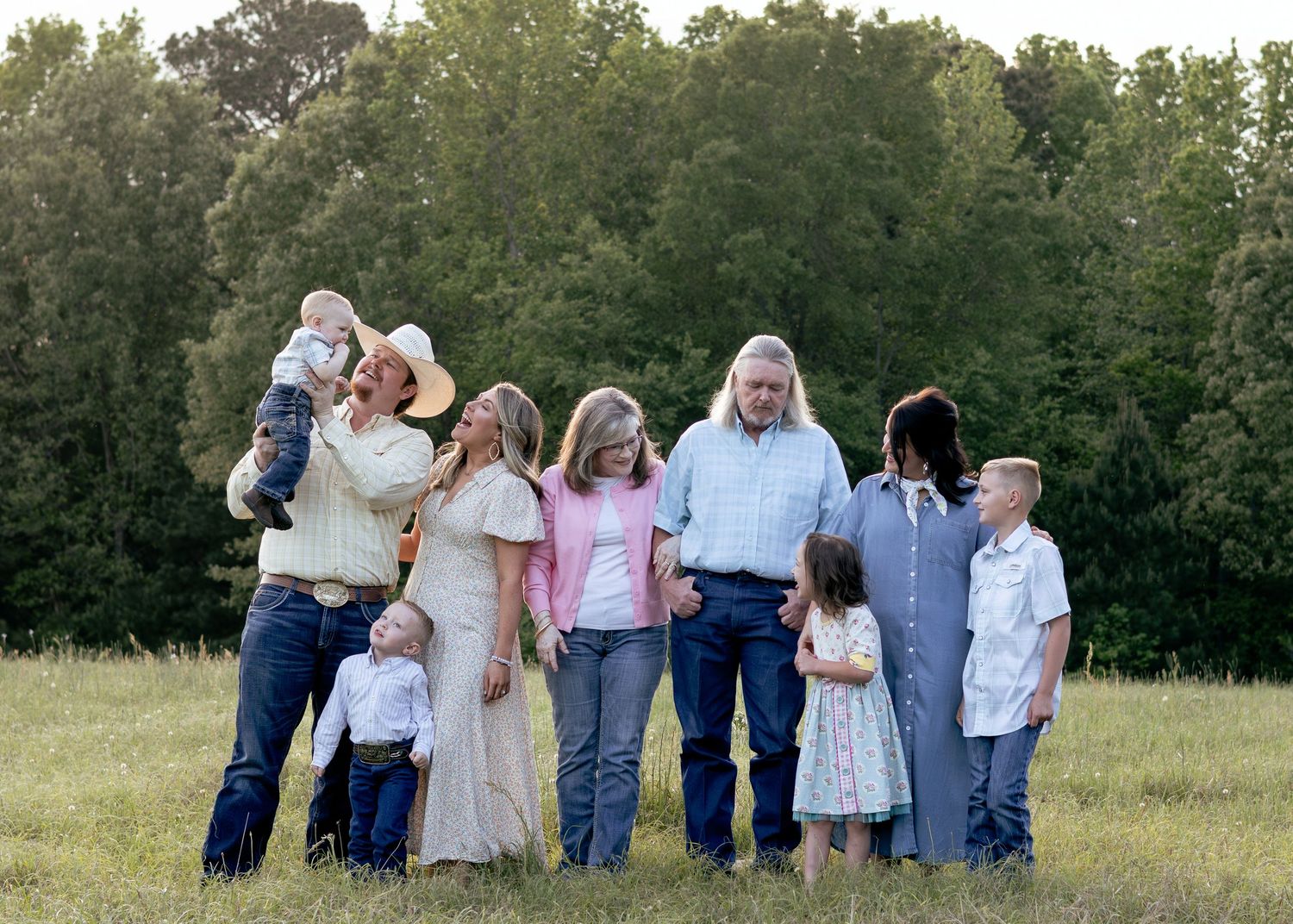 Large family posing together in a green meadow with tall trees, including children and adults in casual country attire.
