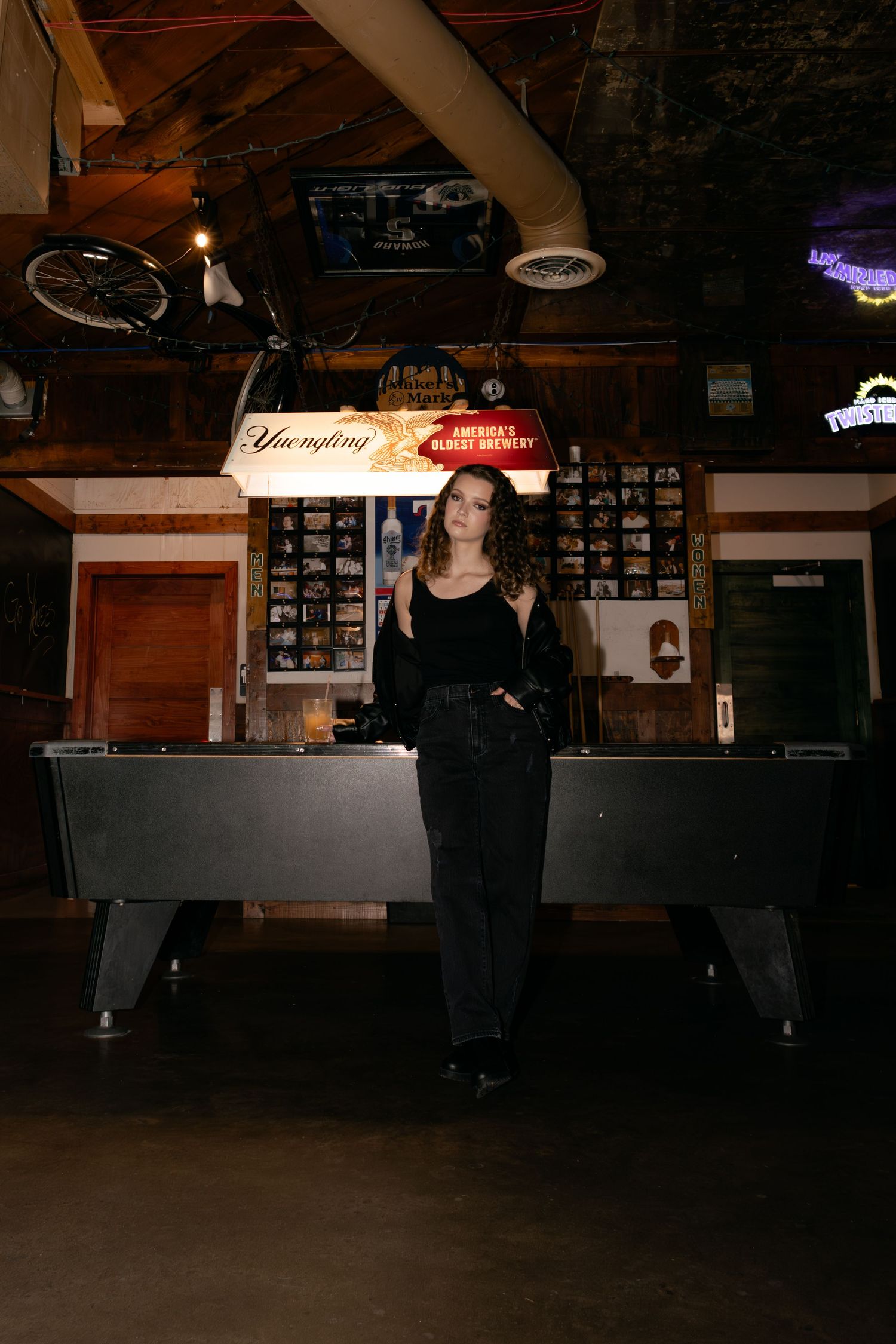 Woman in black outfit stands before a pool table in a rustic bar with neon signs and vintage decor.