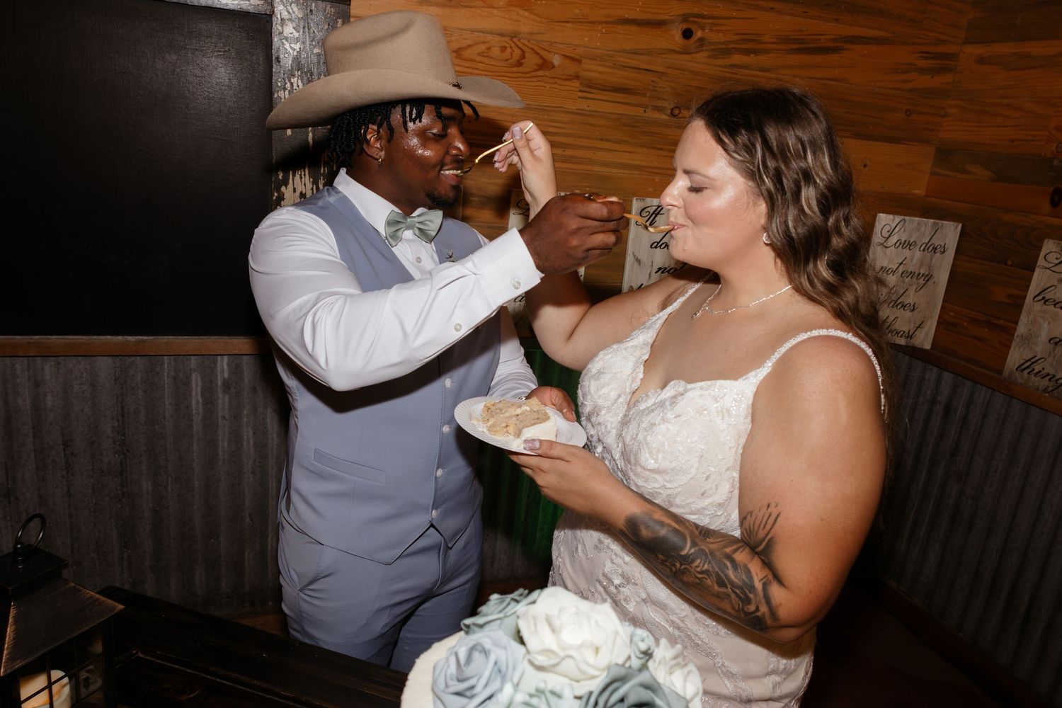 Bride and groom sharing wedding cake during reception, groom wearing cowboy hat and blue suit.