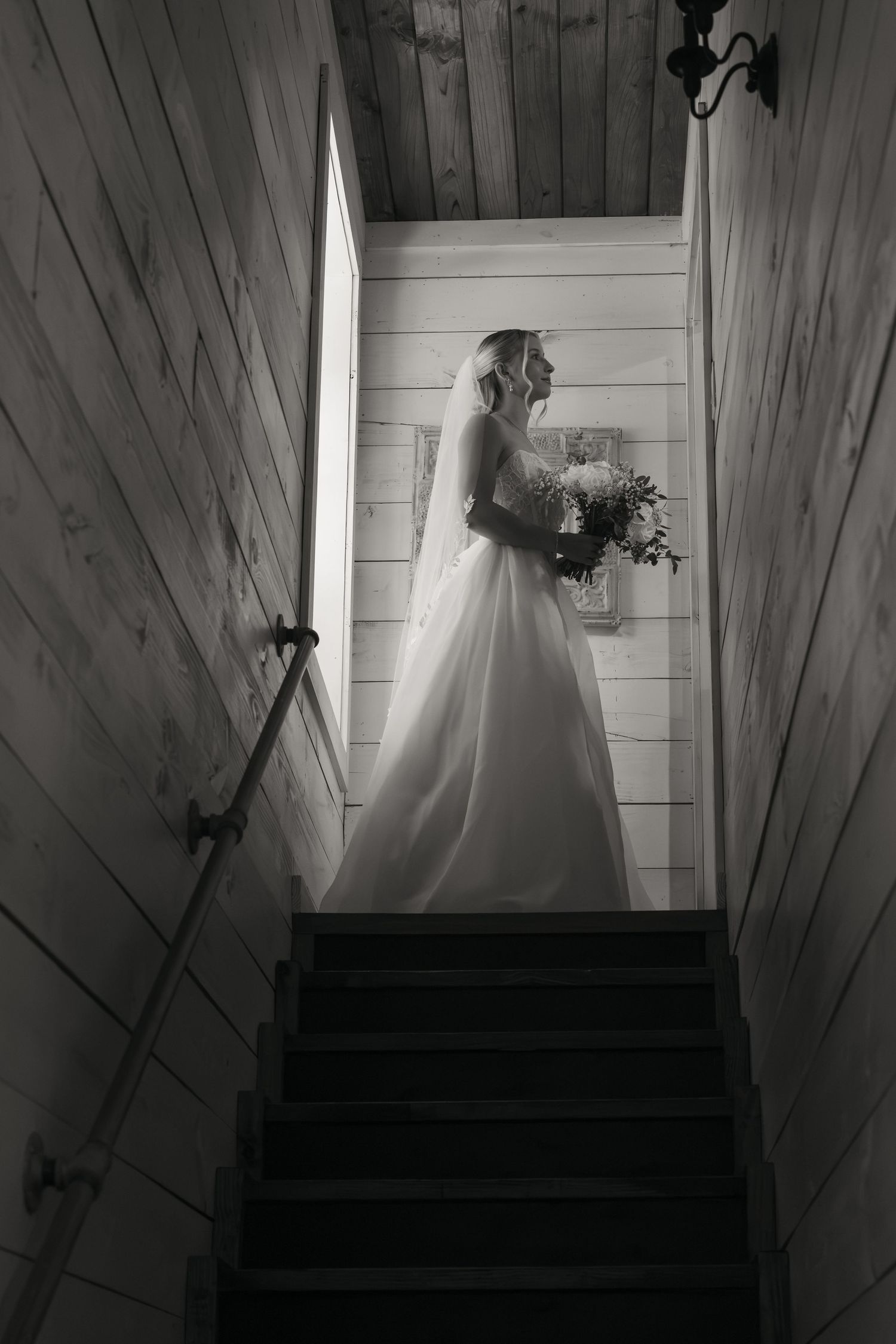 Bride in white gown and veil holding bouquet stands at top of staircase in black and white wedding photo.