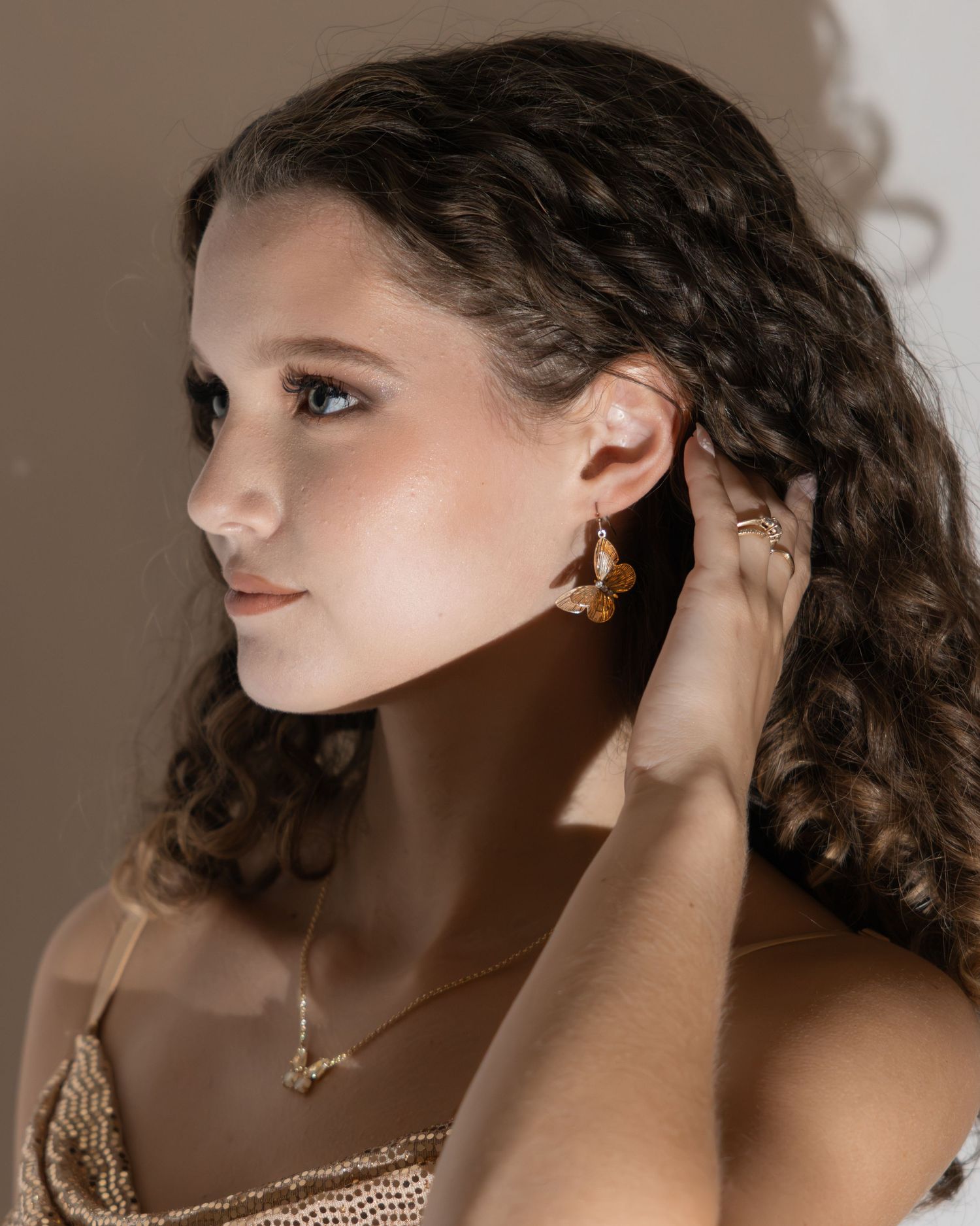 Young woman with curly hair wearing gold butterfly earrings and a delicate necklace in profile view.