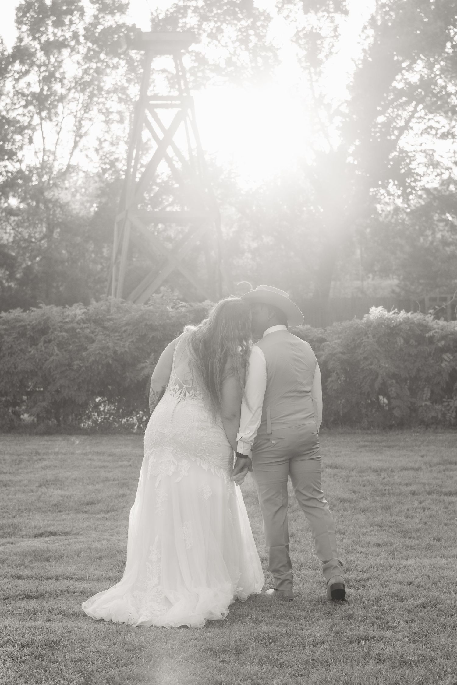 Black and white photo of bride and groom holding hands, walking toward sunlit trees in a romantic outdoor wedding setting.