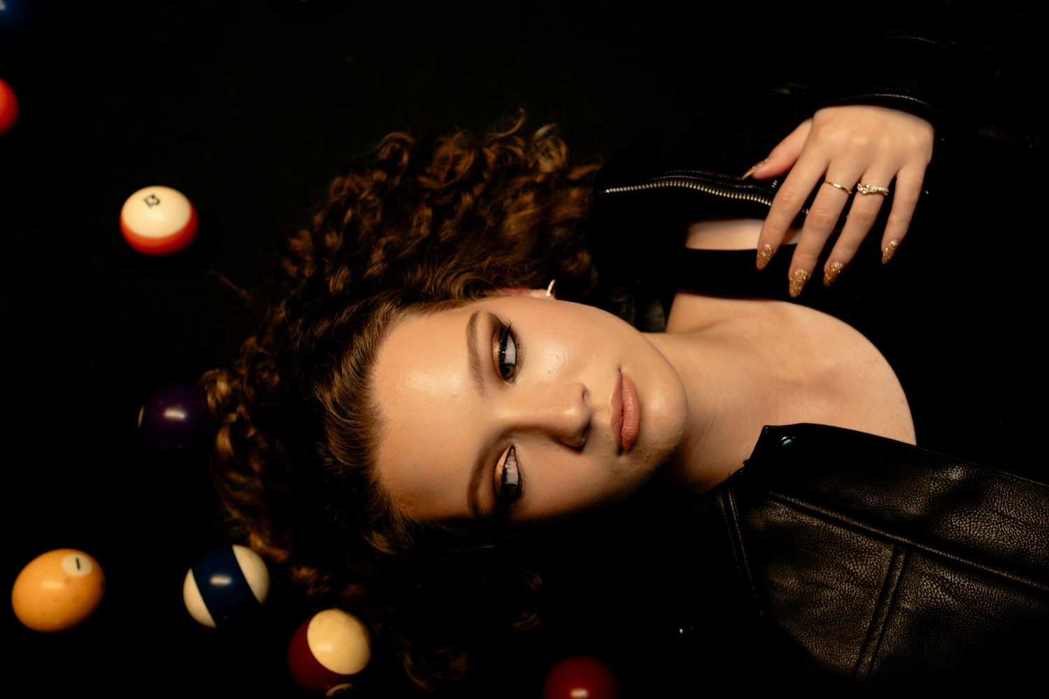 Young woman with curly updo lying among colorful billiard balls and candles, wearing a black leather jacket.