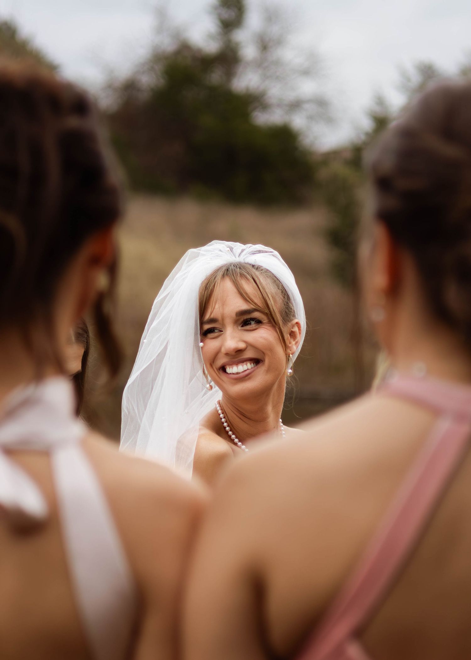 Wedding party scene with bridesmaids in pink dresses gathered around smiling bride wearing white veil.