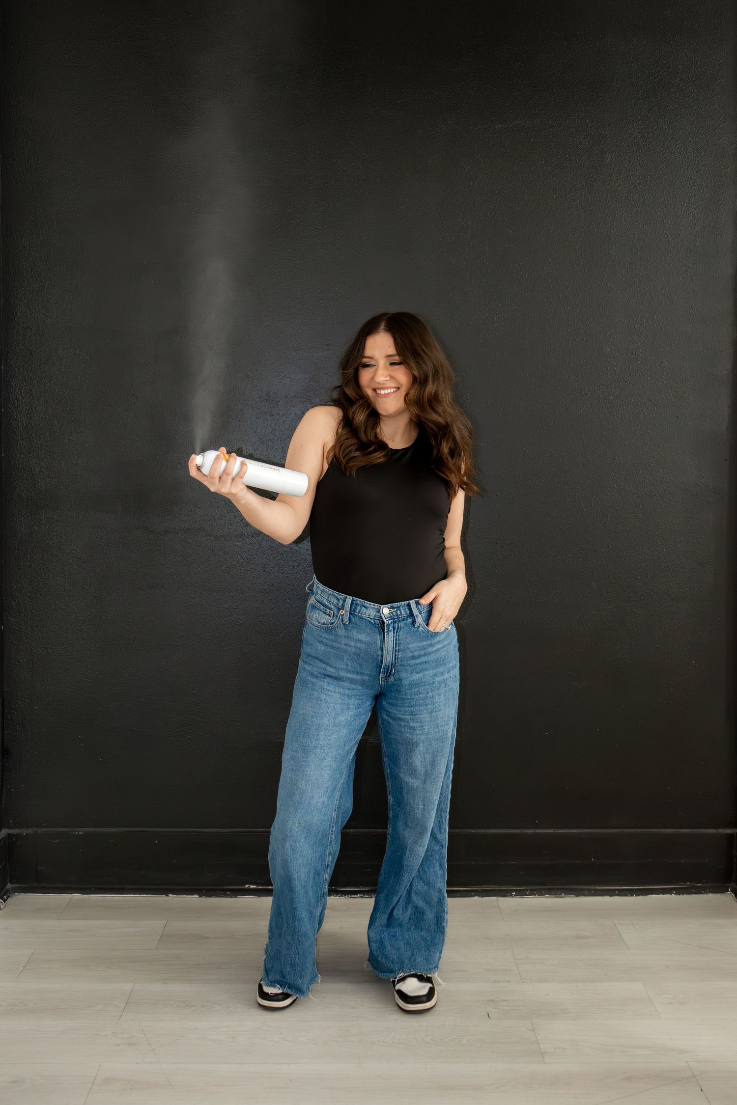 Woman in black tank top and wide-leg jeans spraying hairspray against dark wall.
