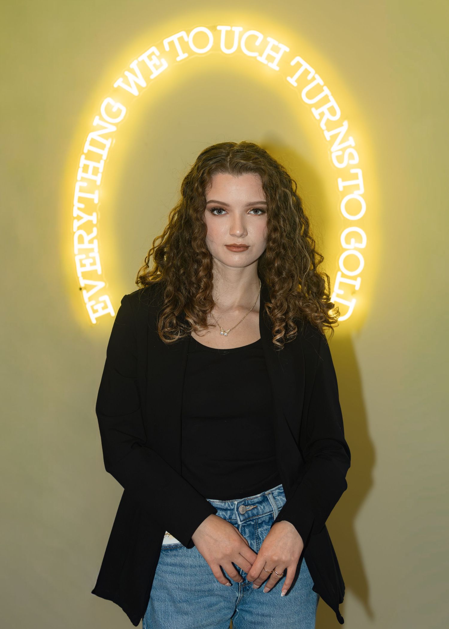 Young woman with curly hair in black blazer and jeans posing before a neon sign reading Everything We Touch Turns To Gold.