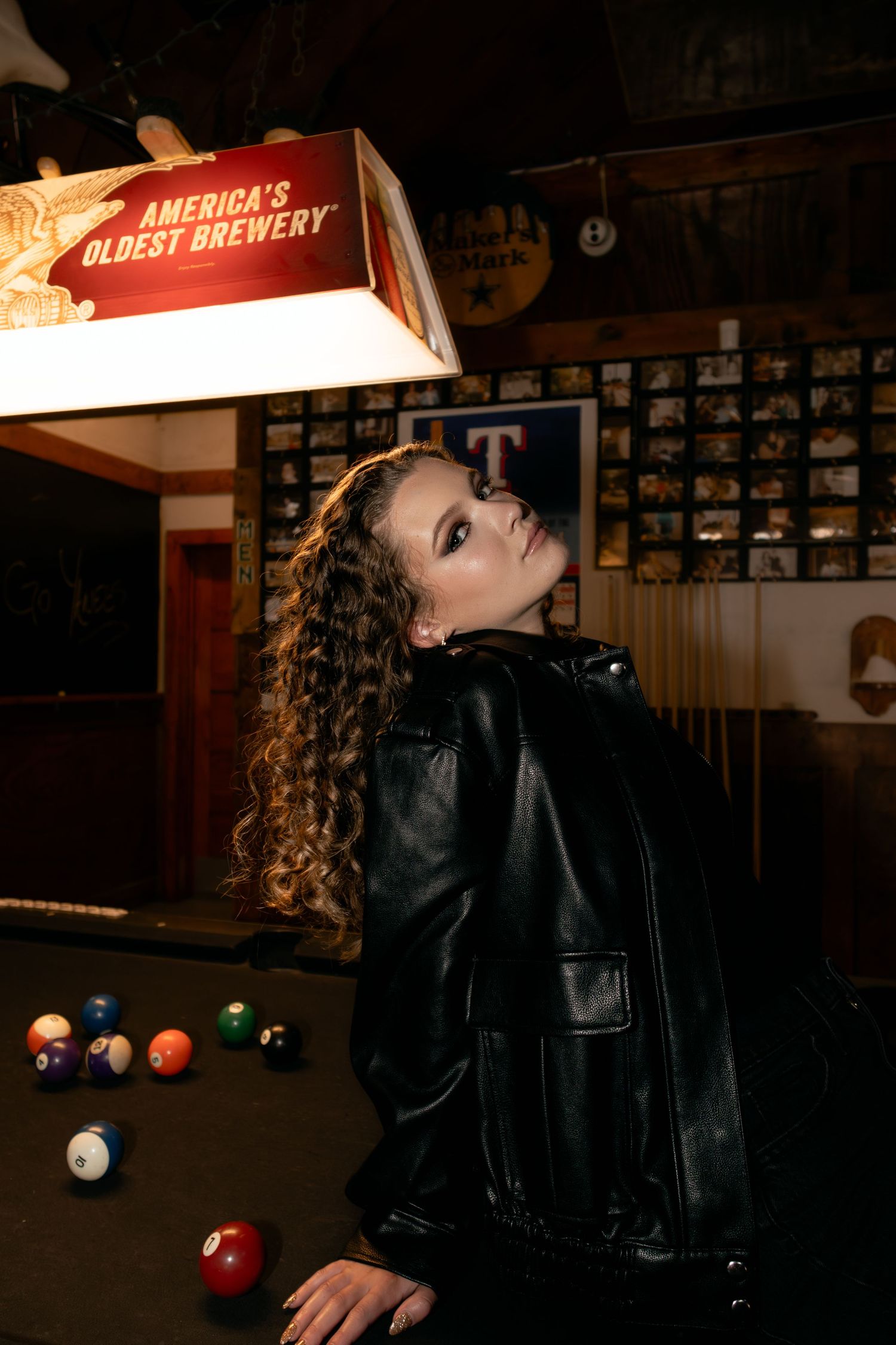 Woman in black leather jacket poses by pool table under Americas Oldest Brewery light in rustic bar.