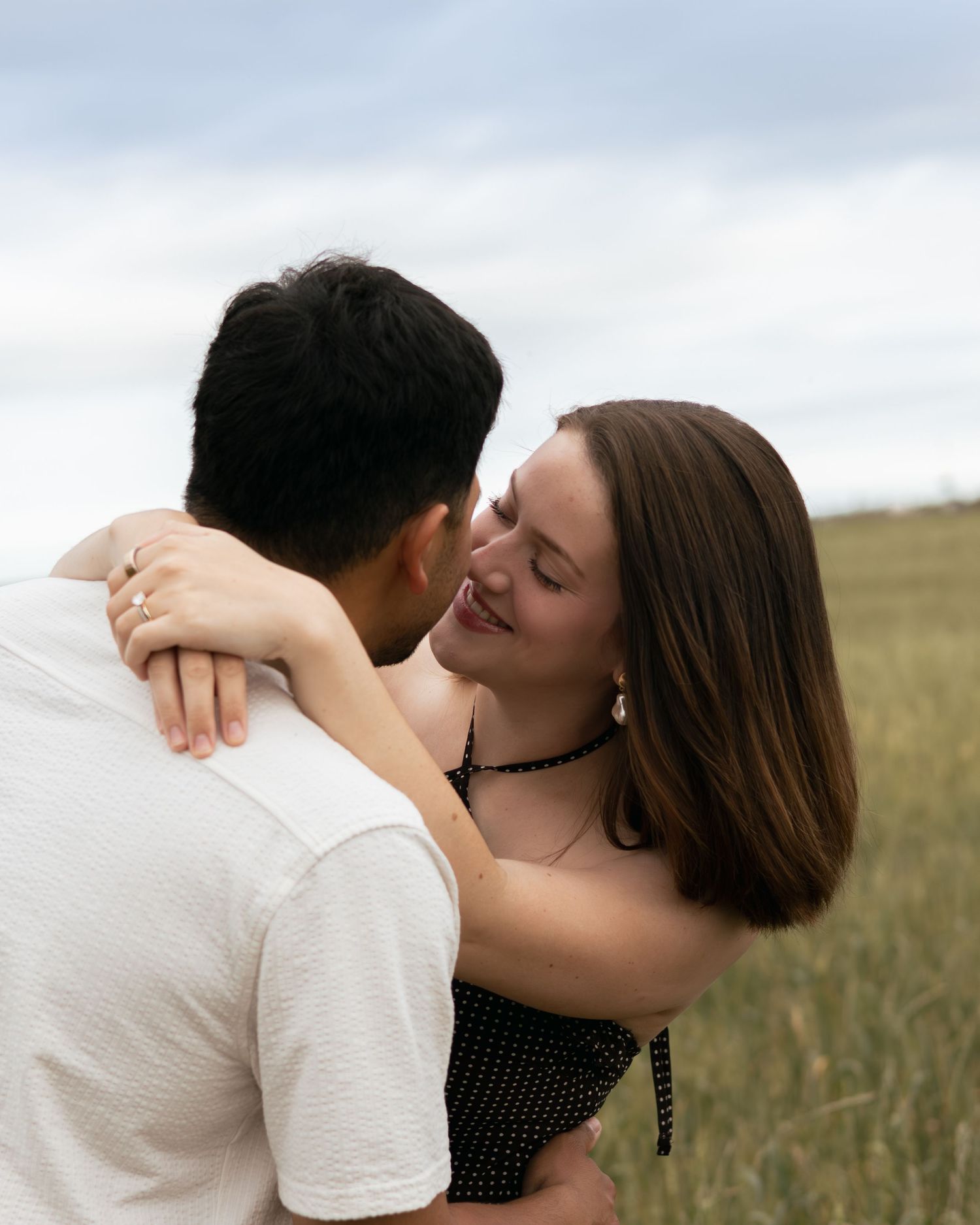 A smiling woman embraces a man in a white shirt in a grassy field, wearing a black dress and ring.