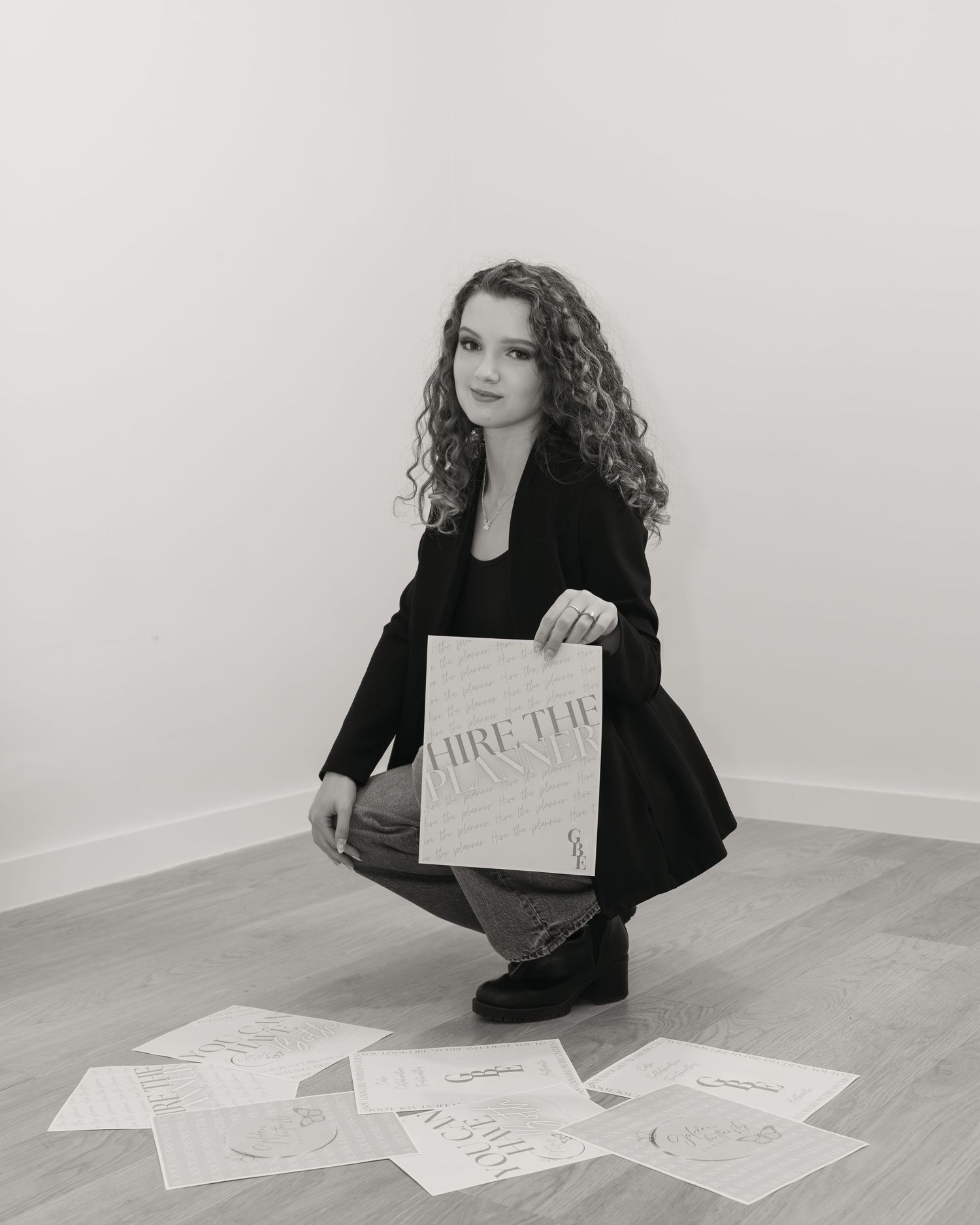 Black and white photo of young woman crouching while holding Hire The Planner book with papers spread on floor.