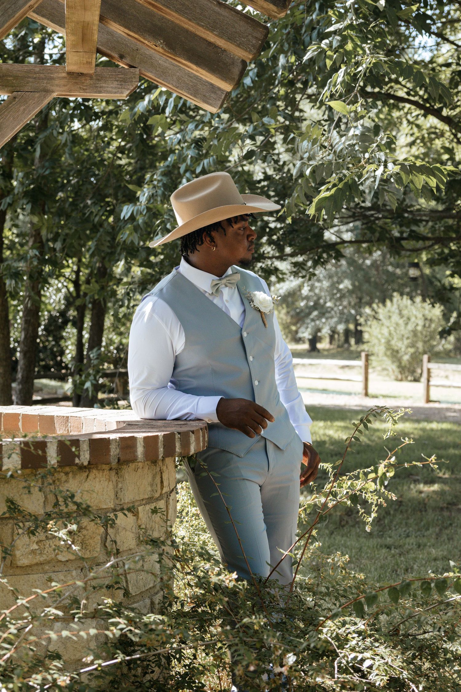 Dapper groom in light blue vest and tan cowboy hat leaning on brick wall outdoors.
