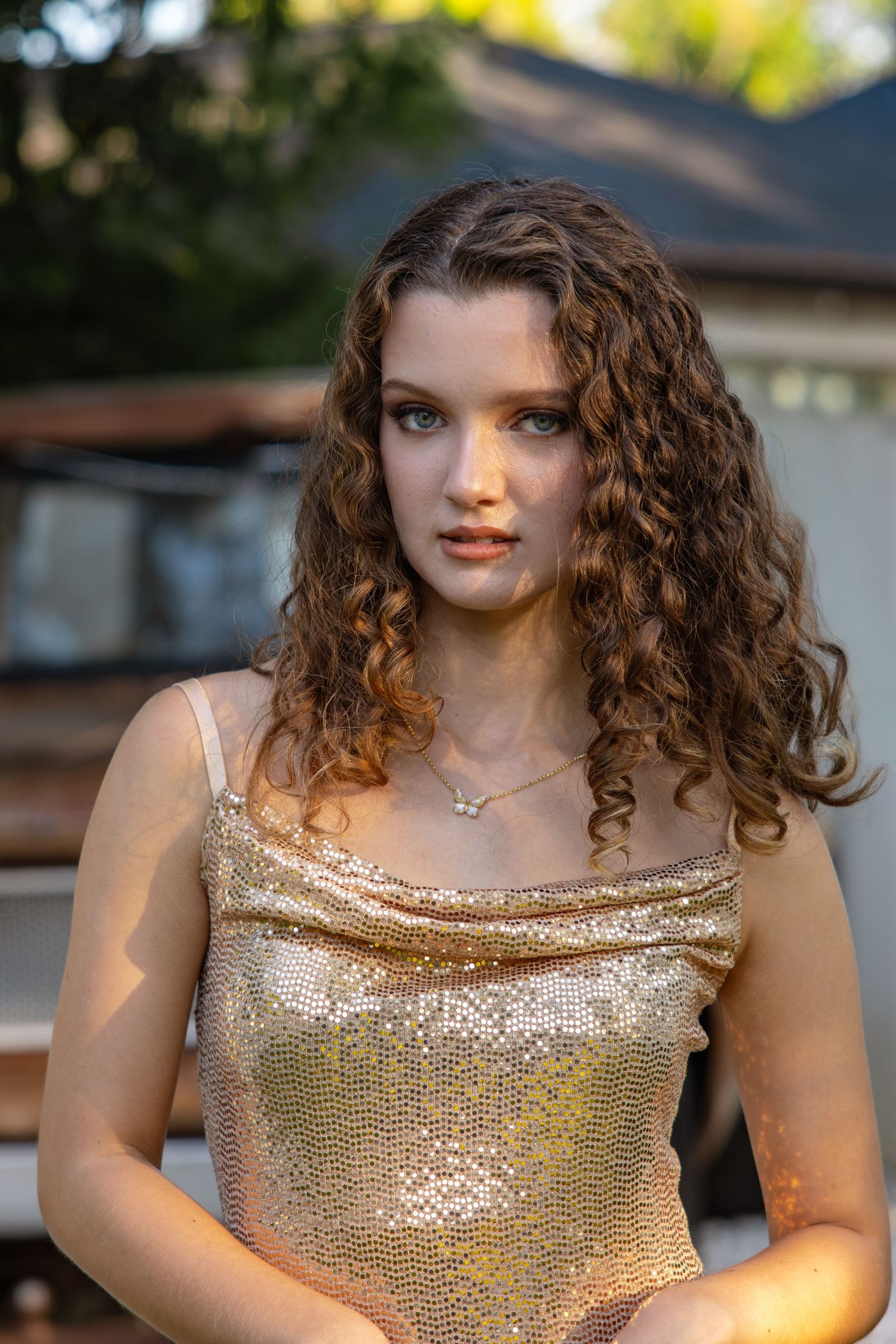 Young woman with curly brown hair wearing gold sequin dress in outdoor golden hour lighting.