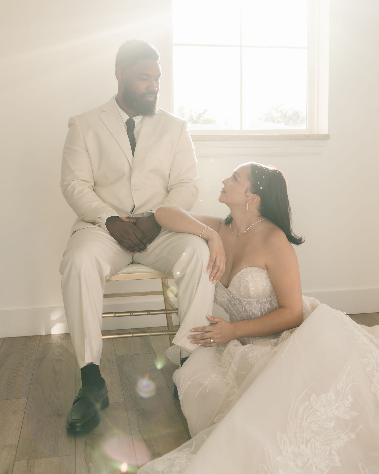 A bride in a white gown sits on the floor gazing up at her groom in a cream suit, bathed in soft natural light.