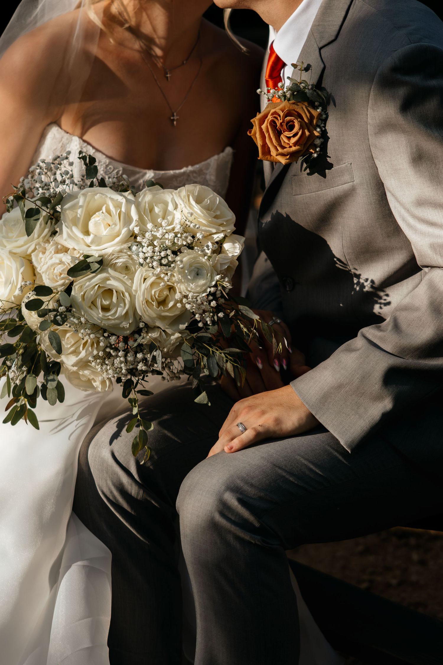 Close-up of bride and groom seated together, white rose bouquet and orange boutonniere visible.