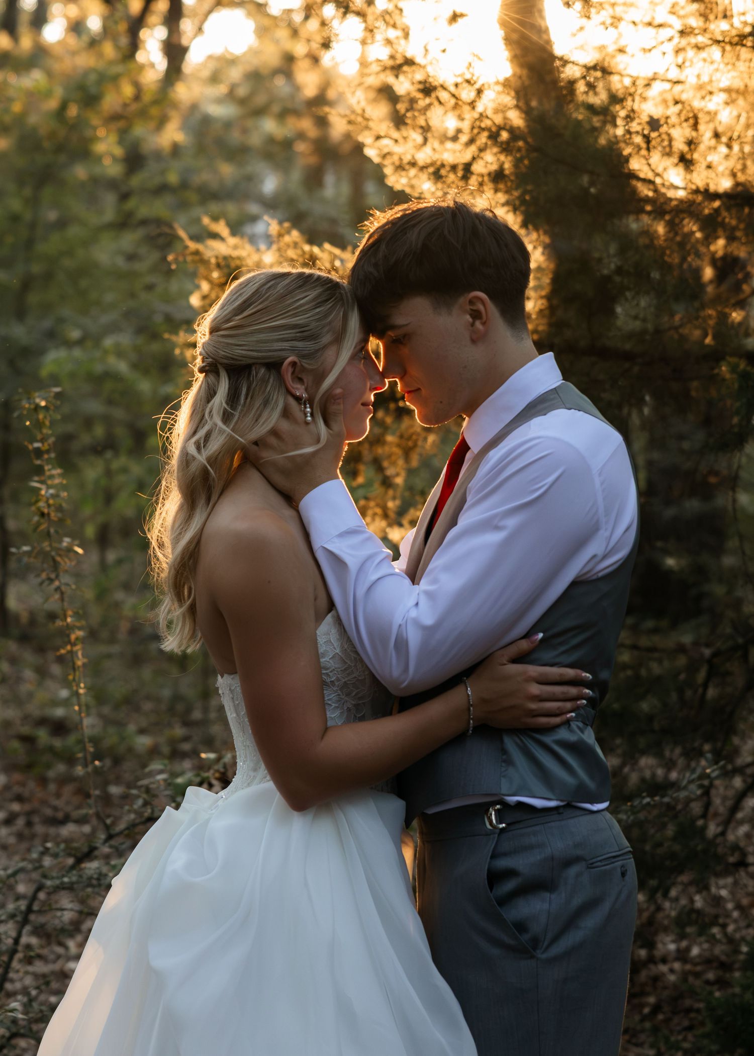 A bride and groom share a romantic forehead touch in a golden sunlit forest, she in white gown, he in gray vest and red tie.