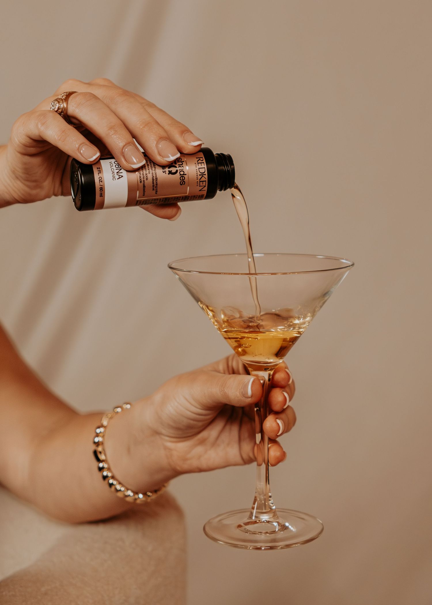 Woman pouring amber liquid from small bottle into elegant martini glass, wearing gold jewelry.