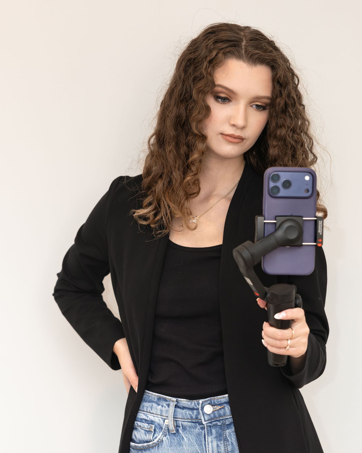 Young woman with curly brown hair holding a smartphone gimbal stabilizer, wearing black blazer and jeans.