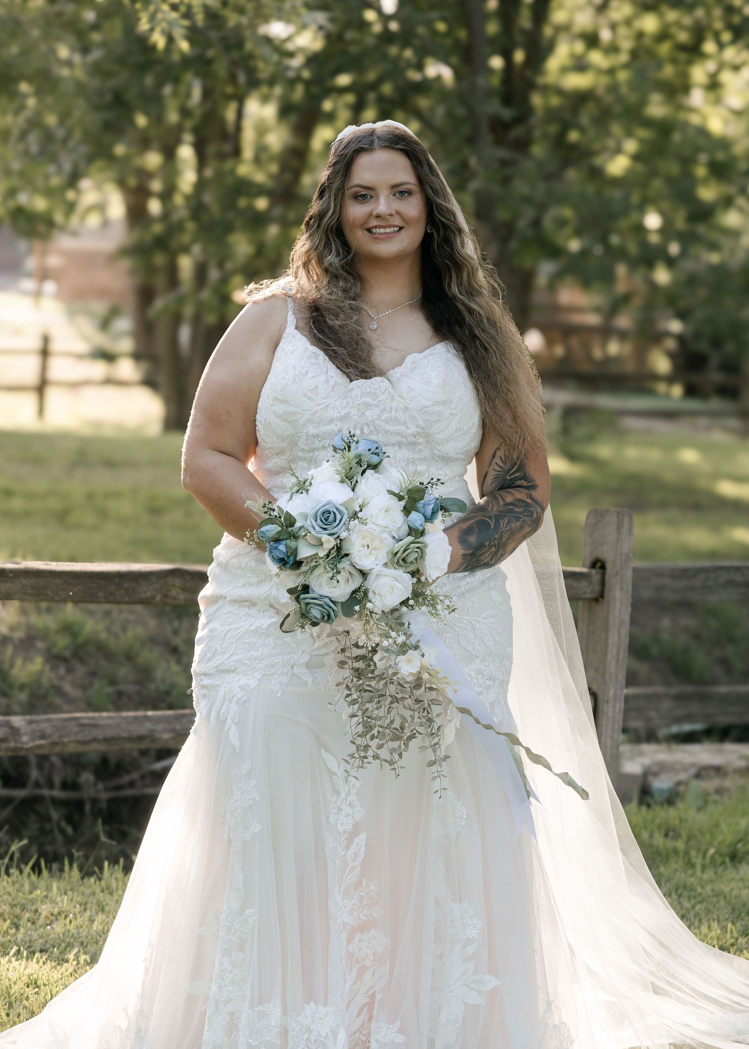 Bride in lace mermaid wedding gown holding blue and white bouquet outdoors by wooden fence.