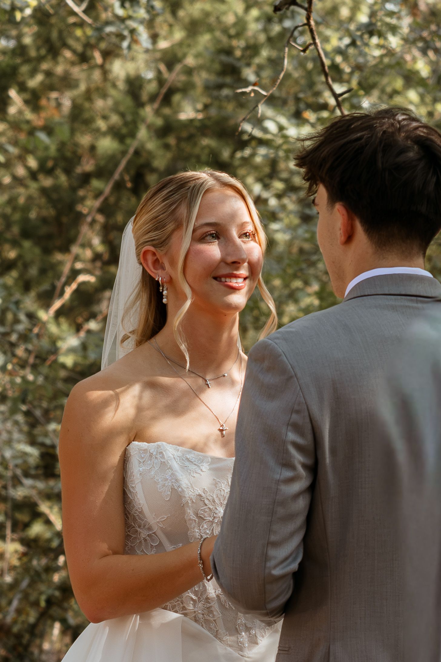 Smiling bride in white lace gown and veil holds hands with groom in gray suit during outdoor wedding ceremony.