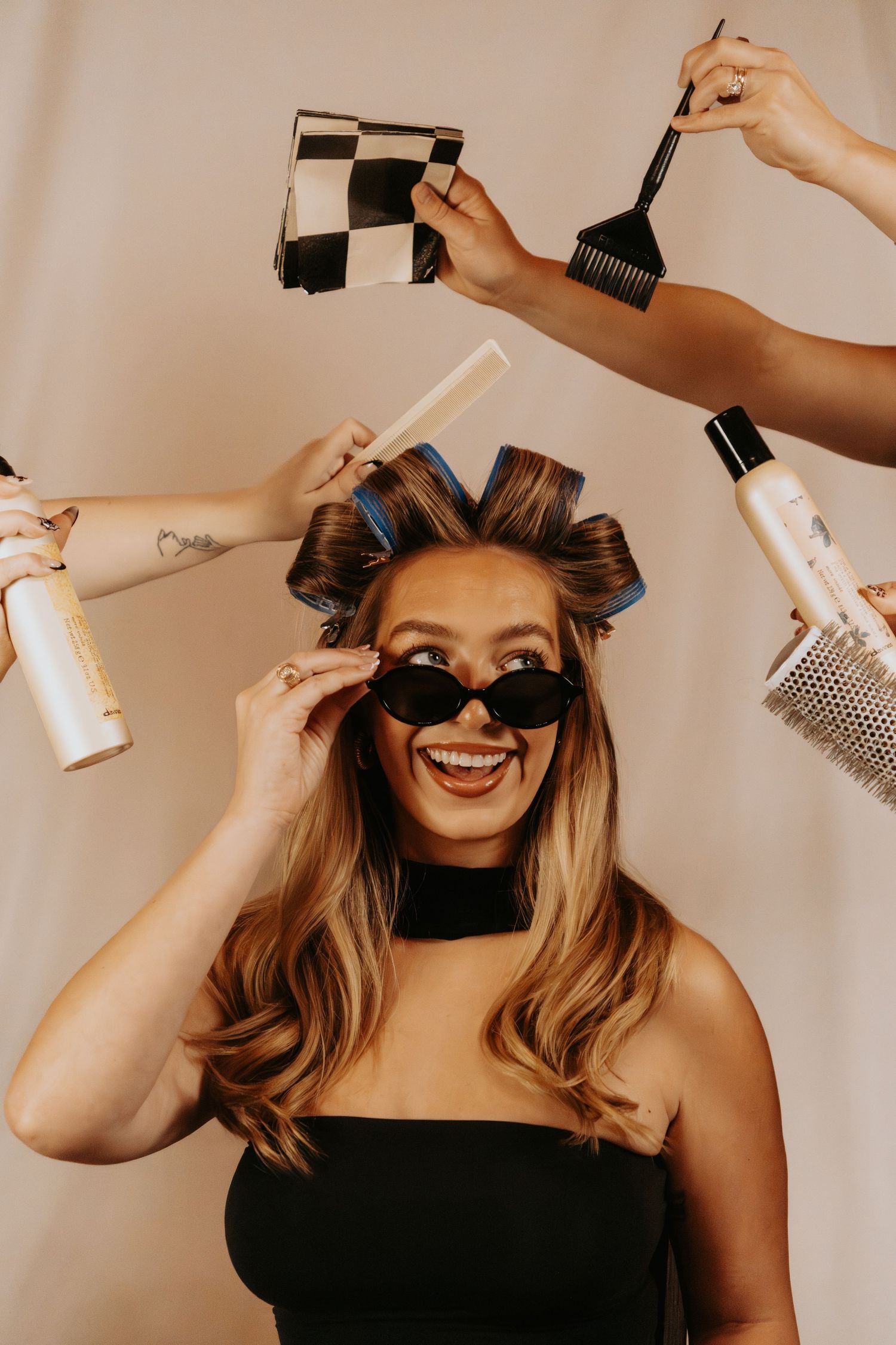 Playful beauty styling session showing various hair and makeup tools being held up against warm beige background.