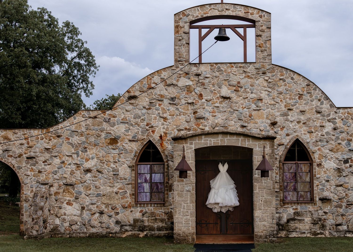 A white wedding dress hangs on a rustic stone chapel door with arched windows and a bell tower under cloudy skies.