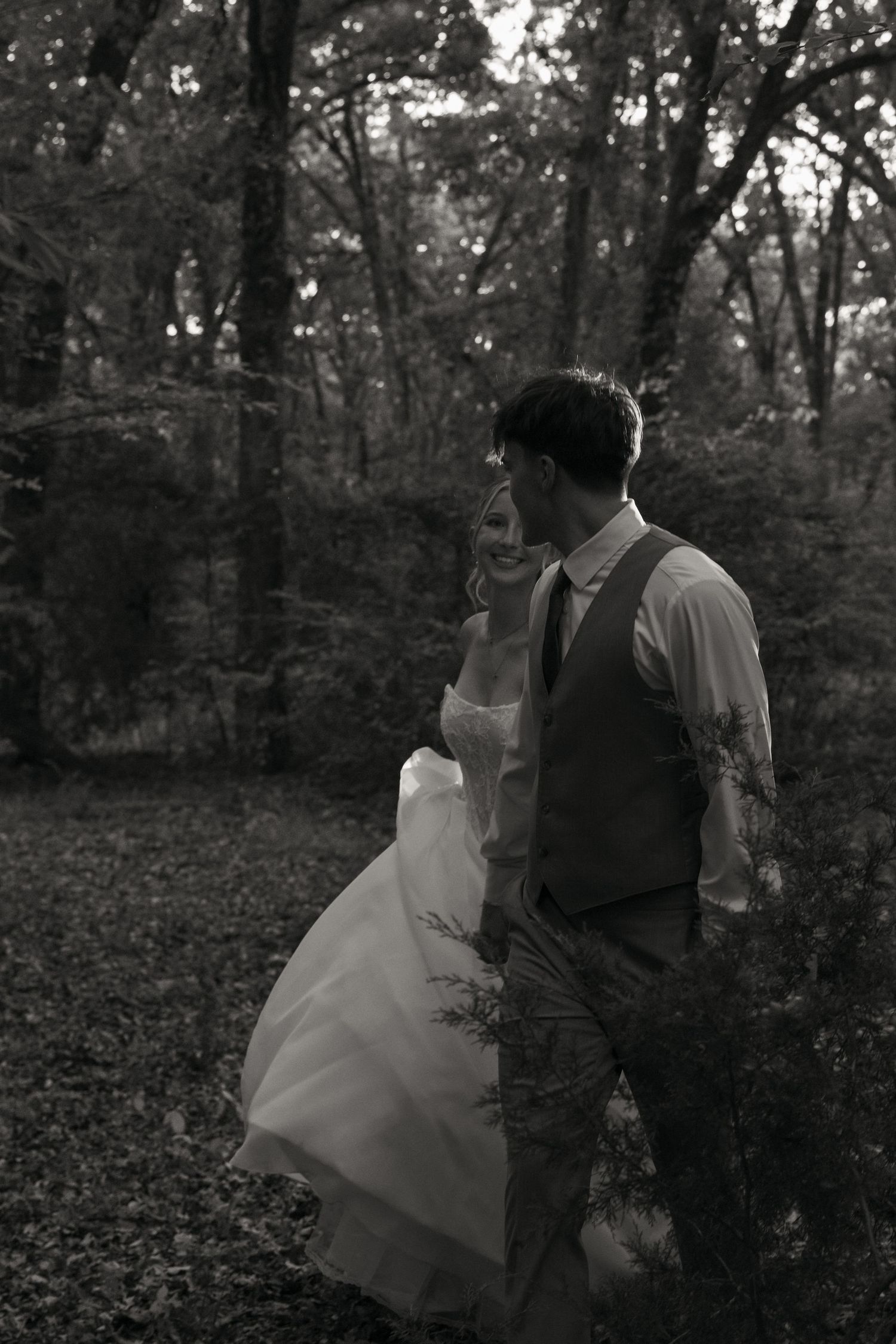 Bride and groom walk hand in hand through a lush forest in an elegant black and white wedding photo.