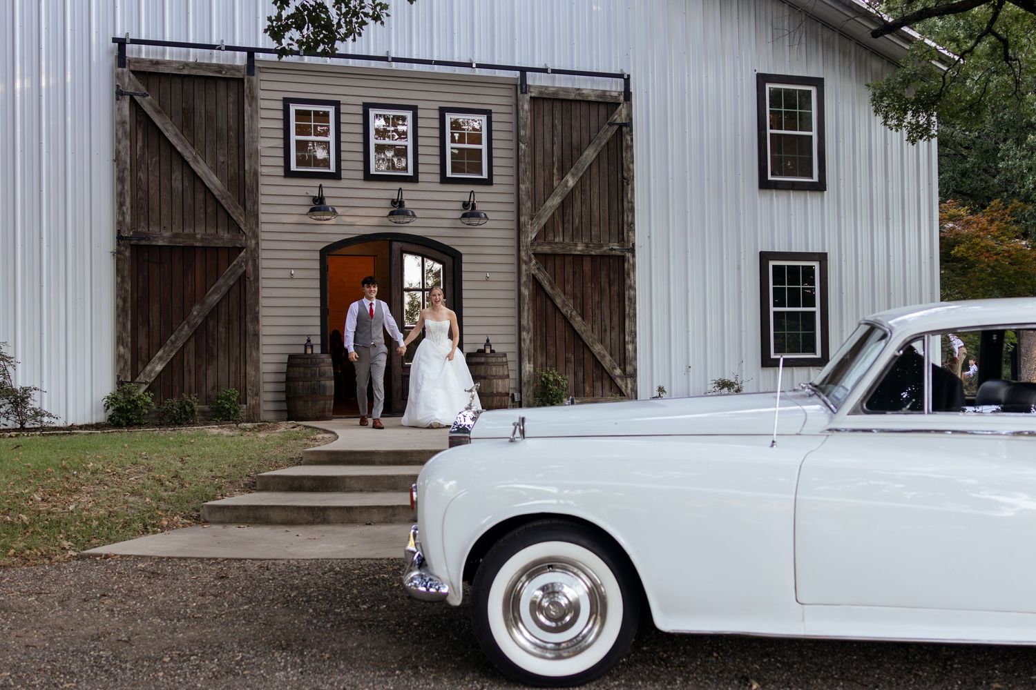 Bride and groom exit a white barn venue beside a classic white vintage car on their wedding day.