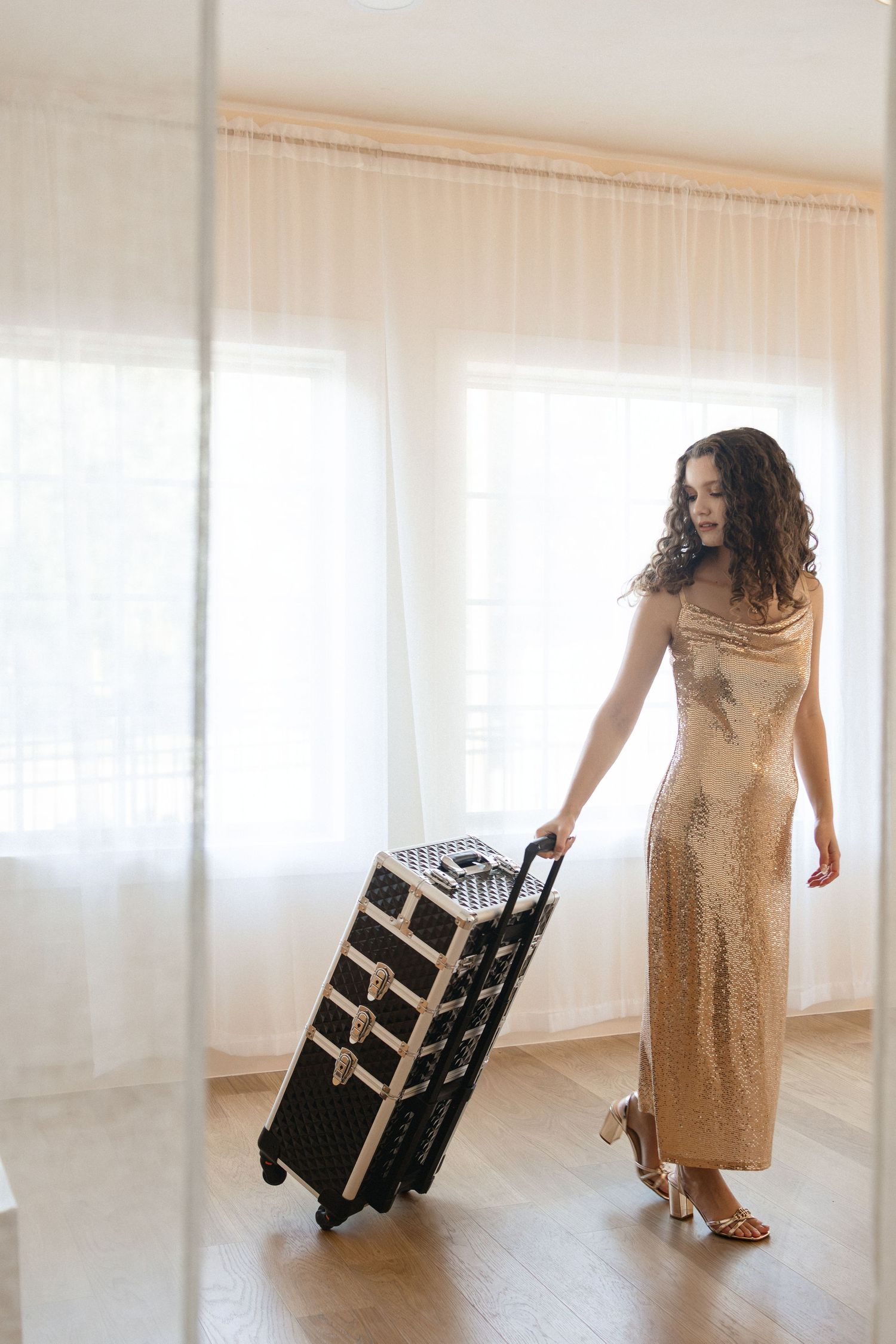 Elegant young woman in a gold sequin gown pulling a professional makeup trolley case indoors.
