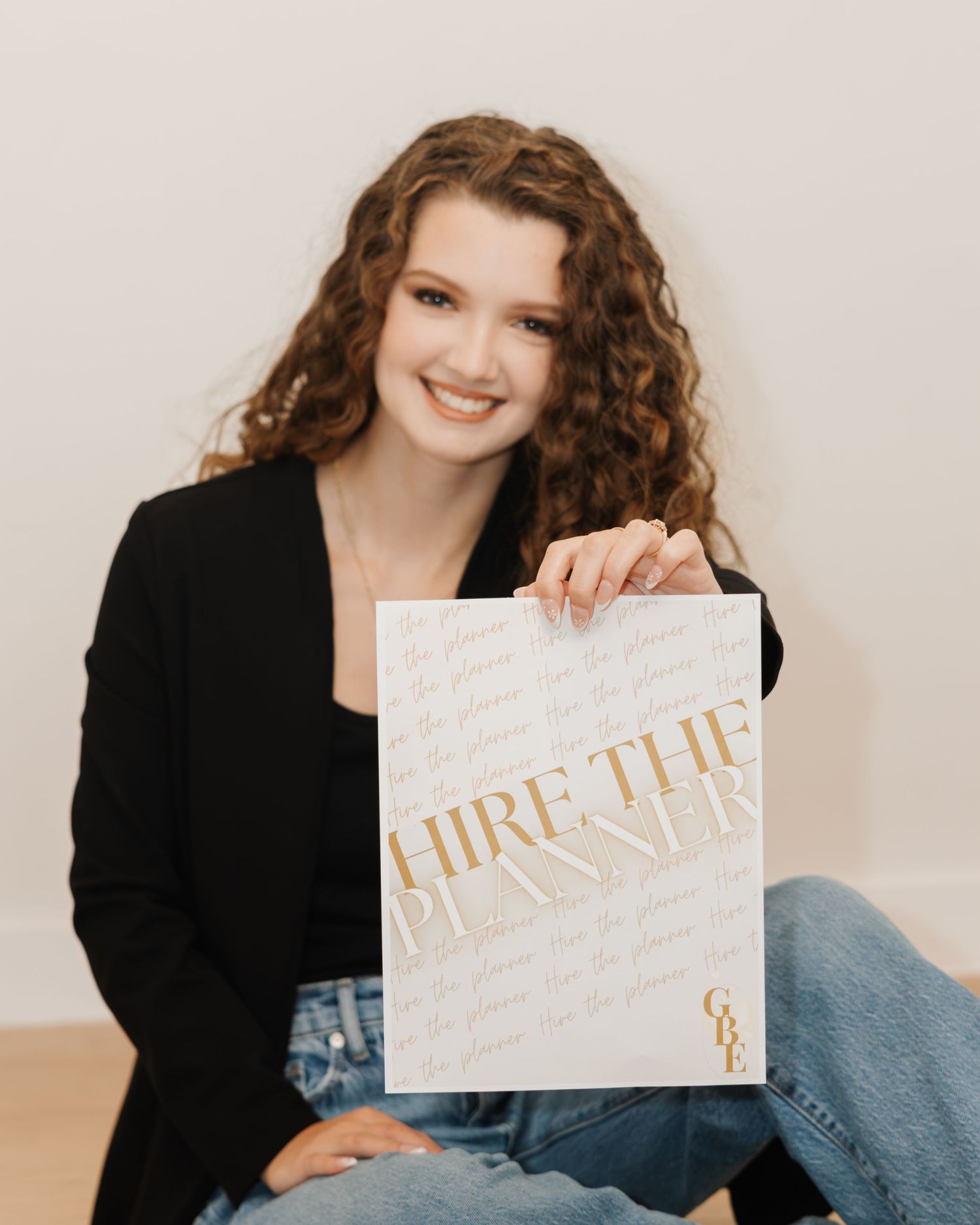 Young woman in black blazer smiling while holding a gold and white Hire The Planner book, seated on floor.