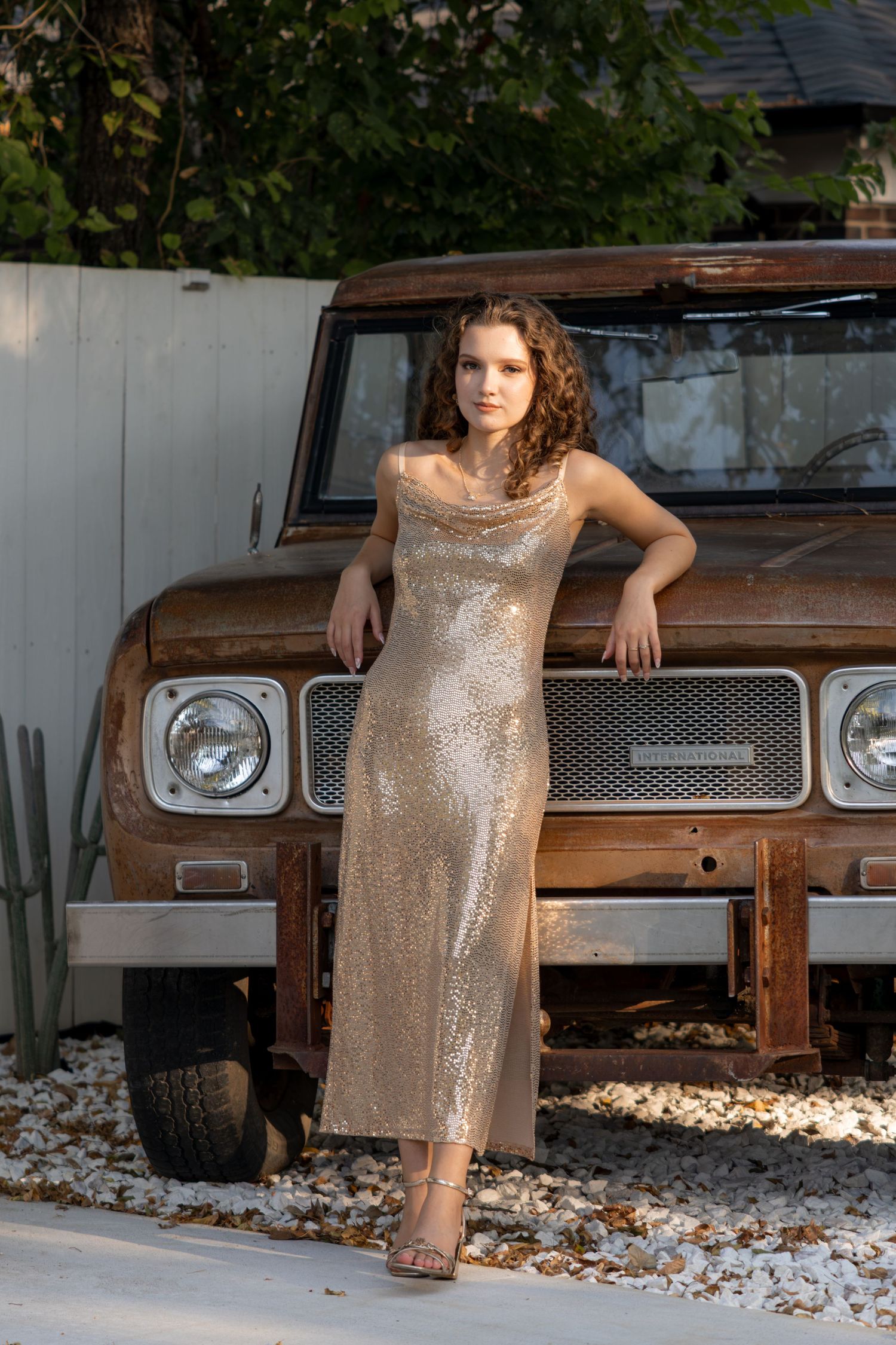 Young woman in a gold sequin maxi dress poses beside a vintage rusty SUV in an outdoor setting.