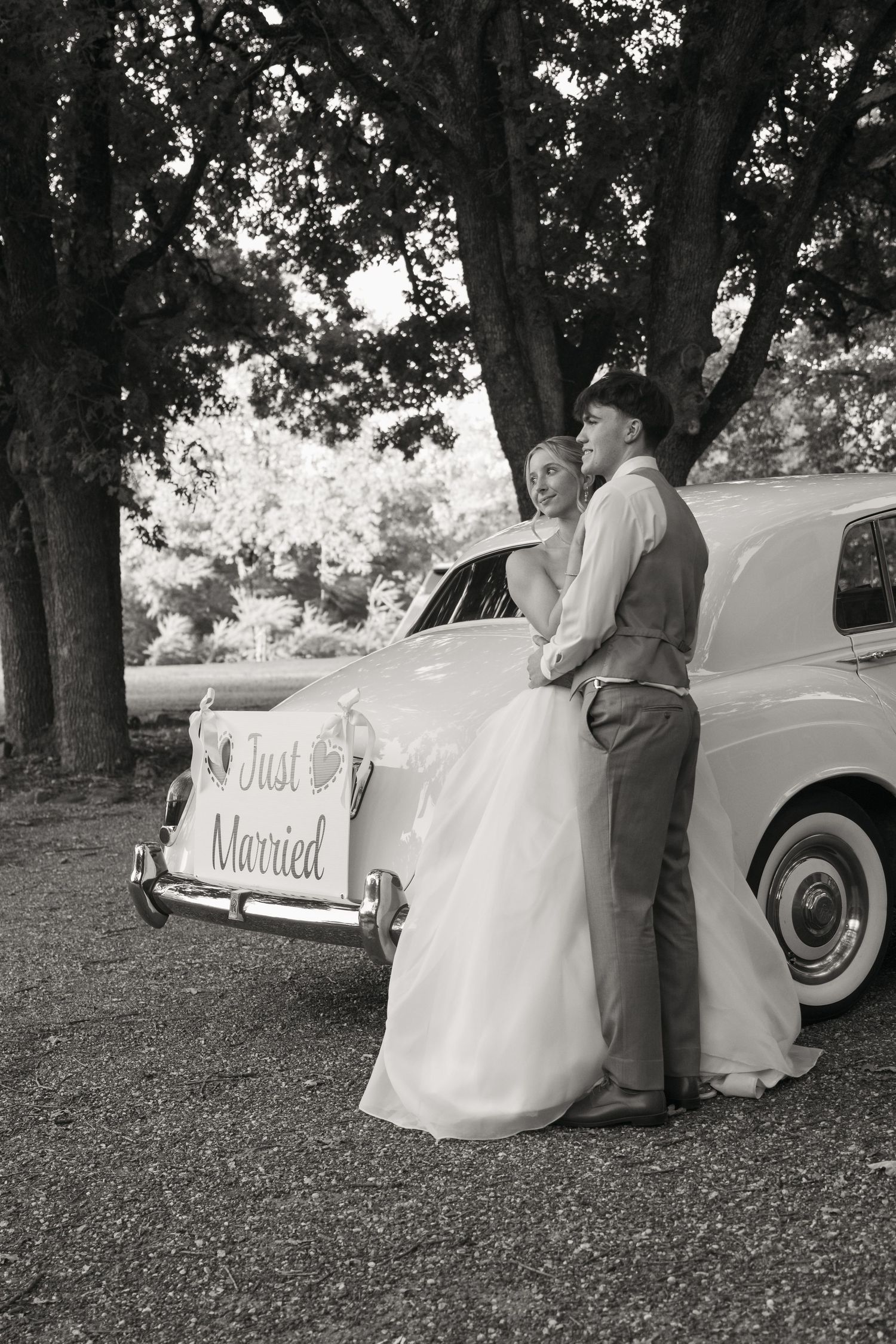 Newlyweds embrace beside vintage Just Married car under trees in classic black and white wedding portrait.