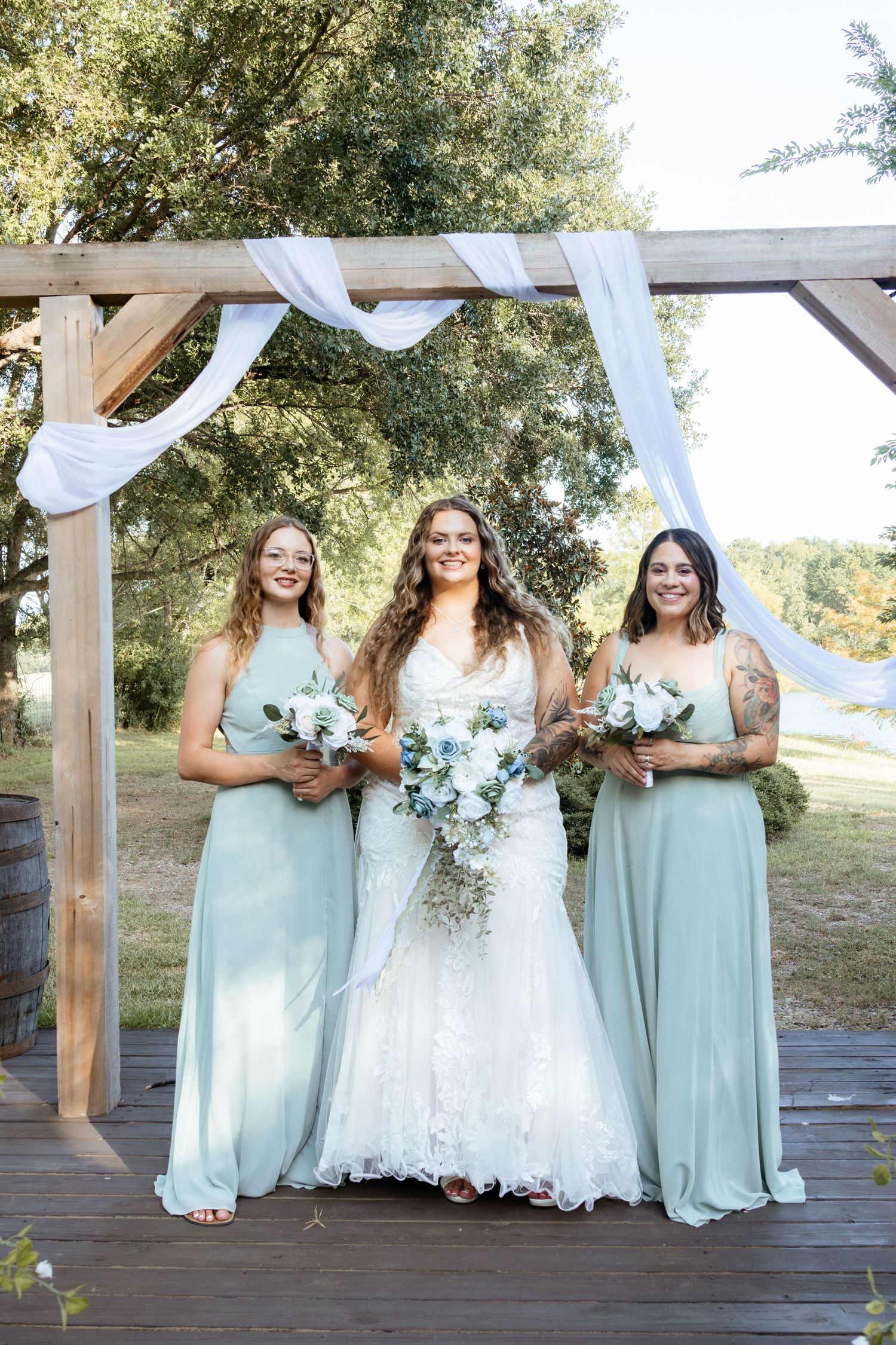 Bride in white lace gown with two bridesmaids in sage green dresses holding bouquets under rustic wooden arch.