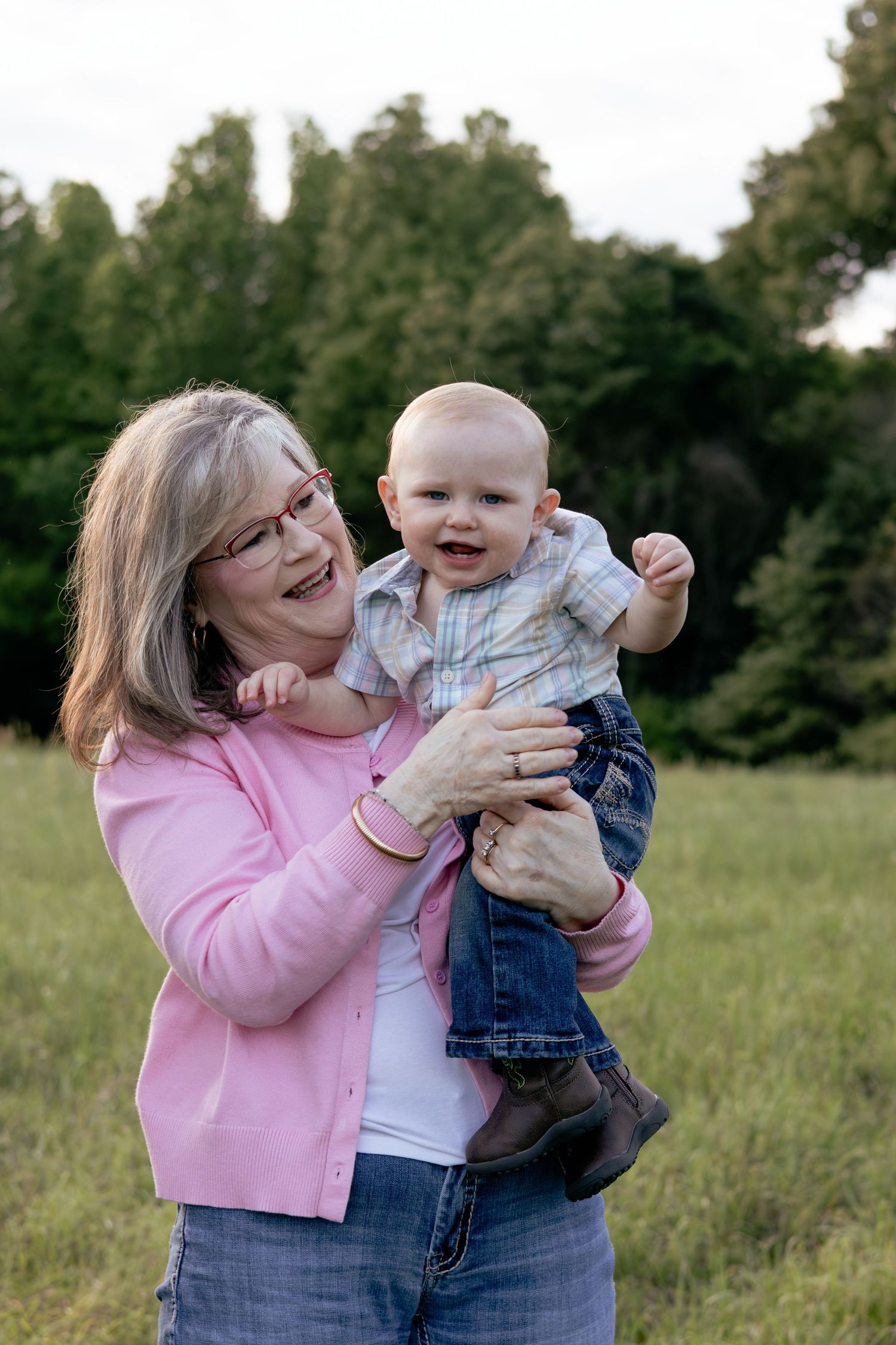 Grandmother in pink cardigan joyfully holds smiling baby outdoors in green field, sharing a happy moment together.