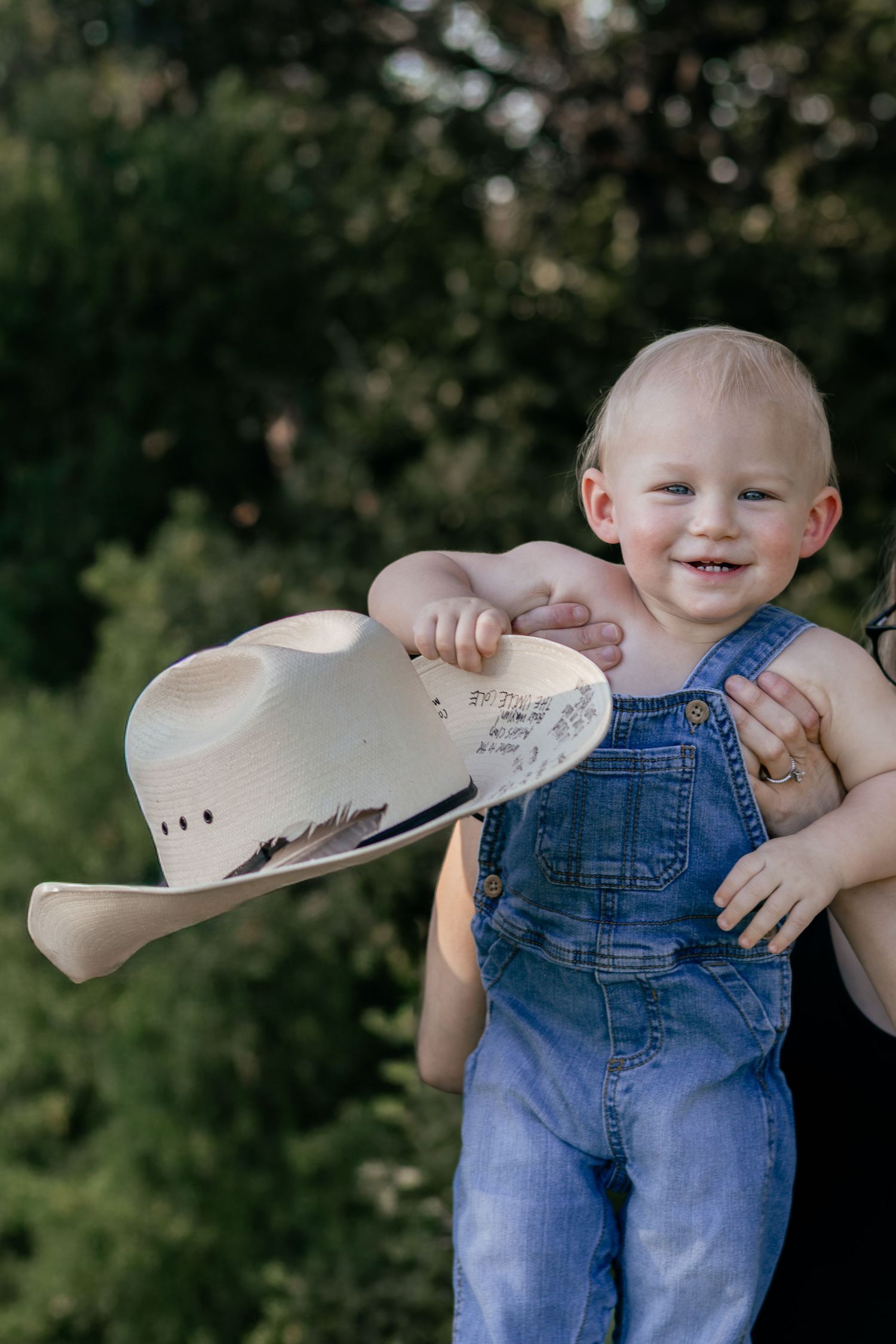 A smiling baby in denim overalls holds a cowboy hat while being carried outside.