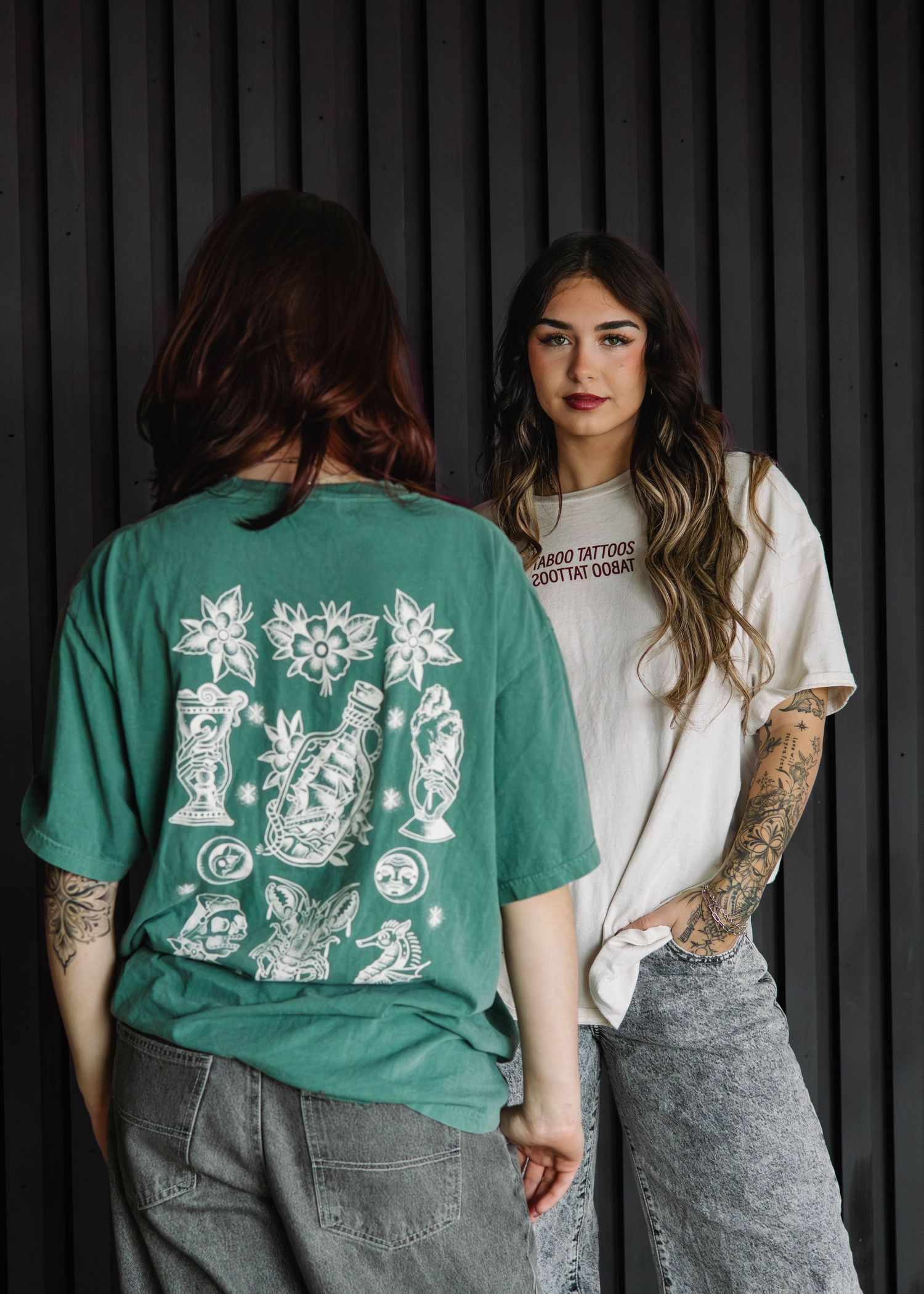 Two women wearing graphic tattoo-themed t-shirts in green and cream, paired with gray jeans, posed against a dark wall.