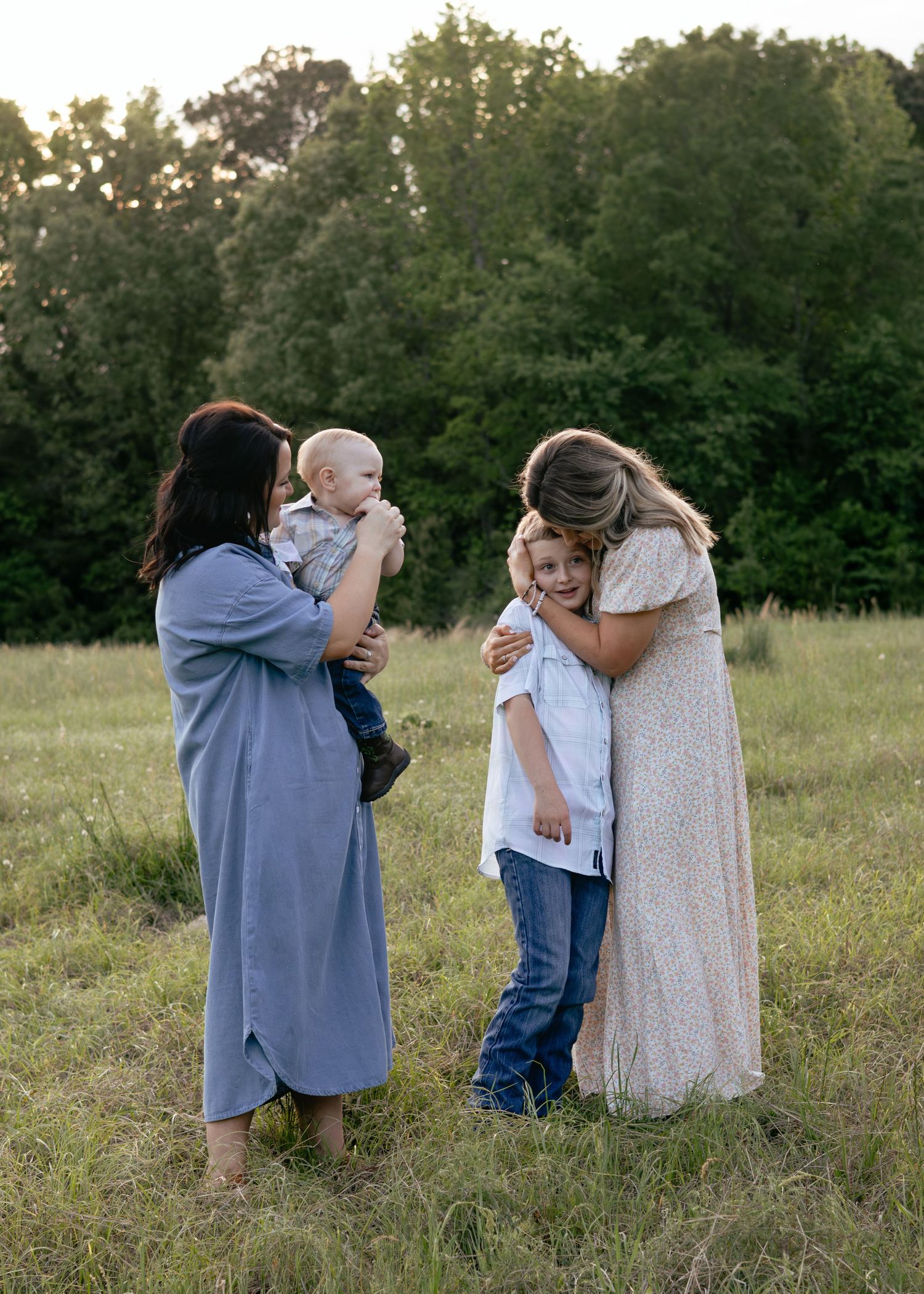 Two mothers with children in a sunny field, one holding a baby, the other hugging an older child, surrounded by green trees.