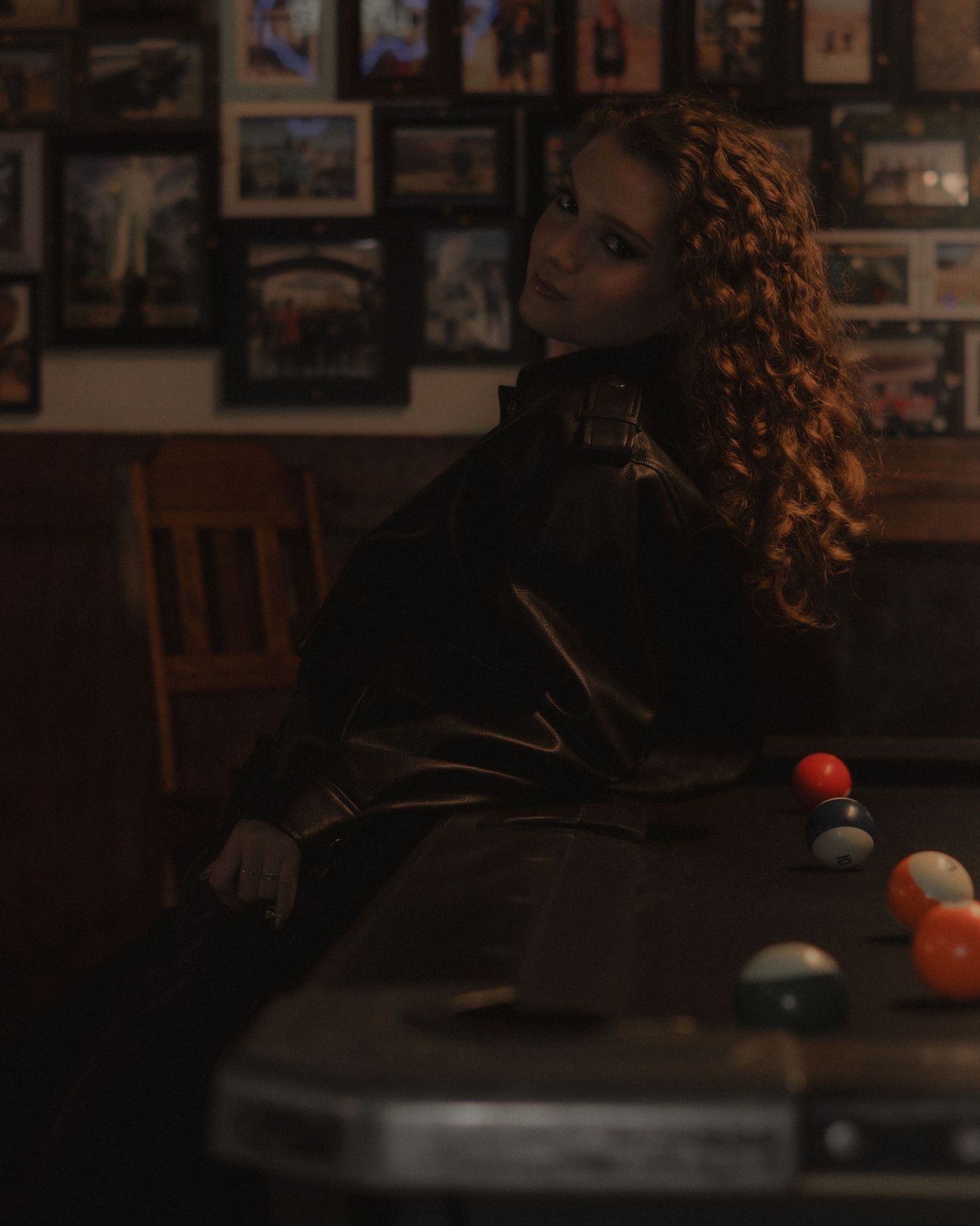 Woman in black leather jacket sits beside a pool table in a dimly lit bar with framed photos.