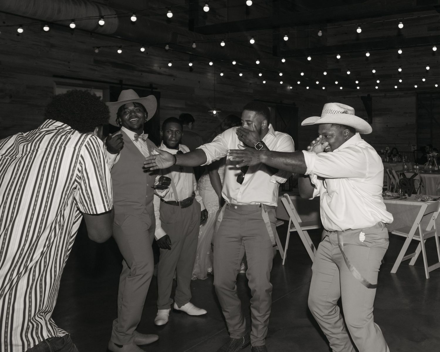Group of men dancing joyfully at a wedding reception in a rustic venue with string lights.