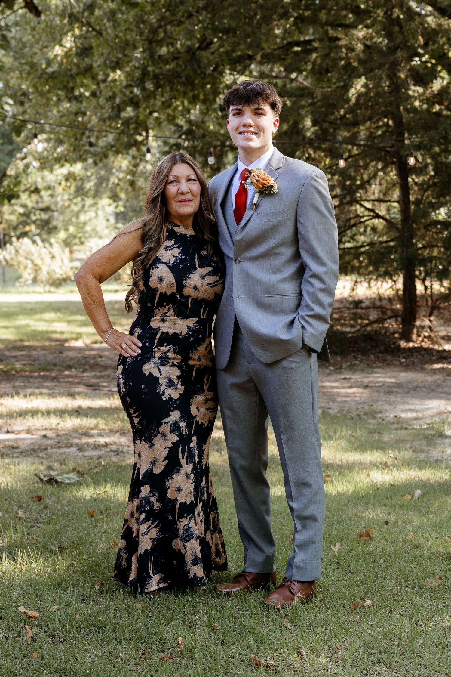 Young man in gray suit with red tie and boutonniere poses with woman in floral dress outdoors.