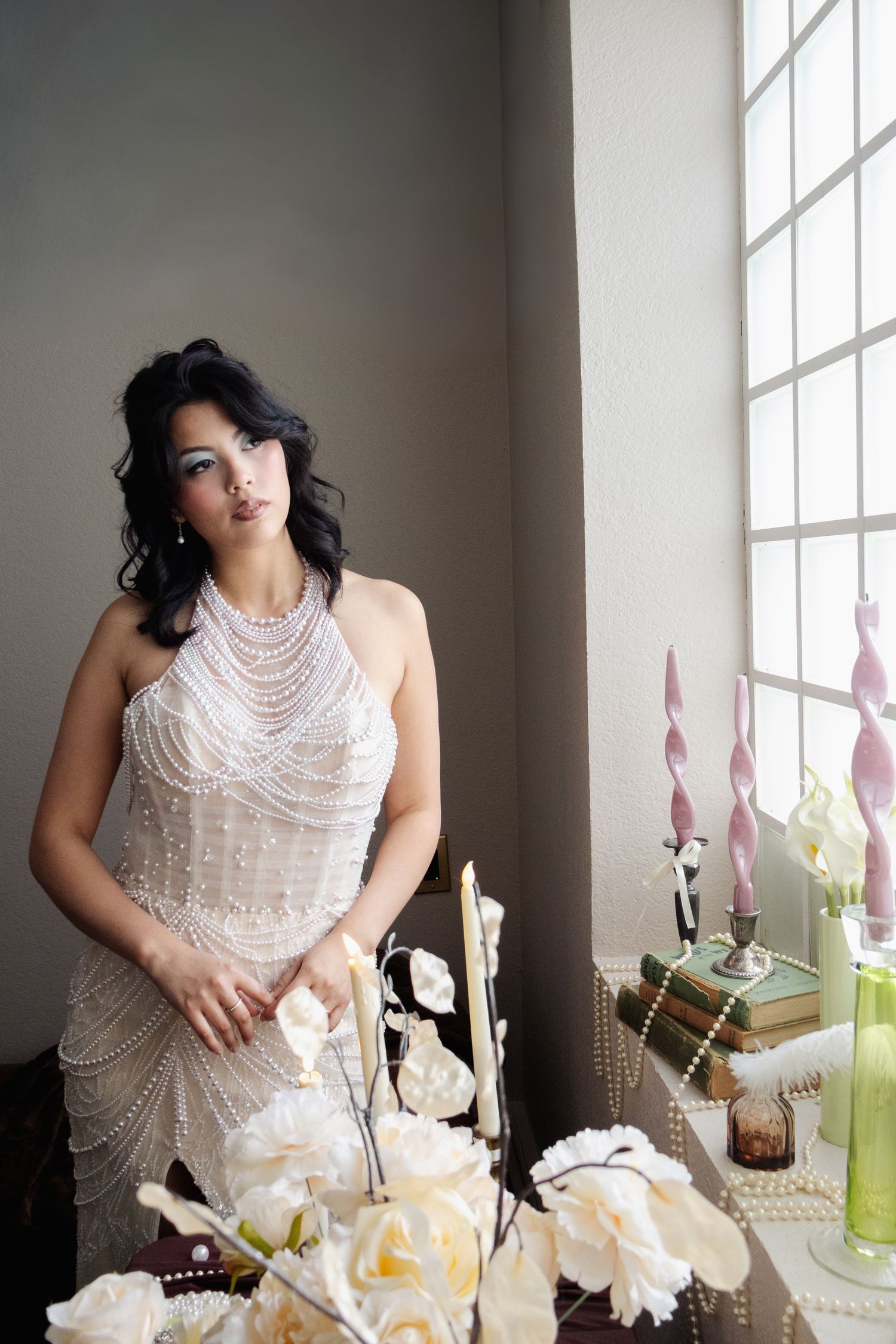 A person in a beaded white gown poses elegantly by a window with white roses and decor in soft natural lighting.