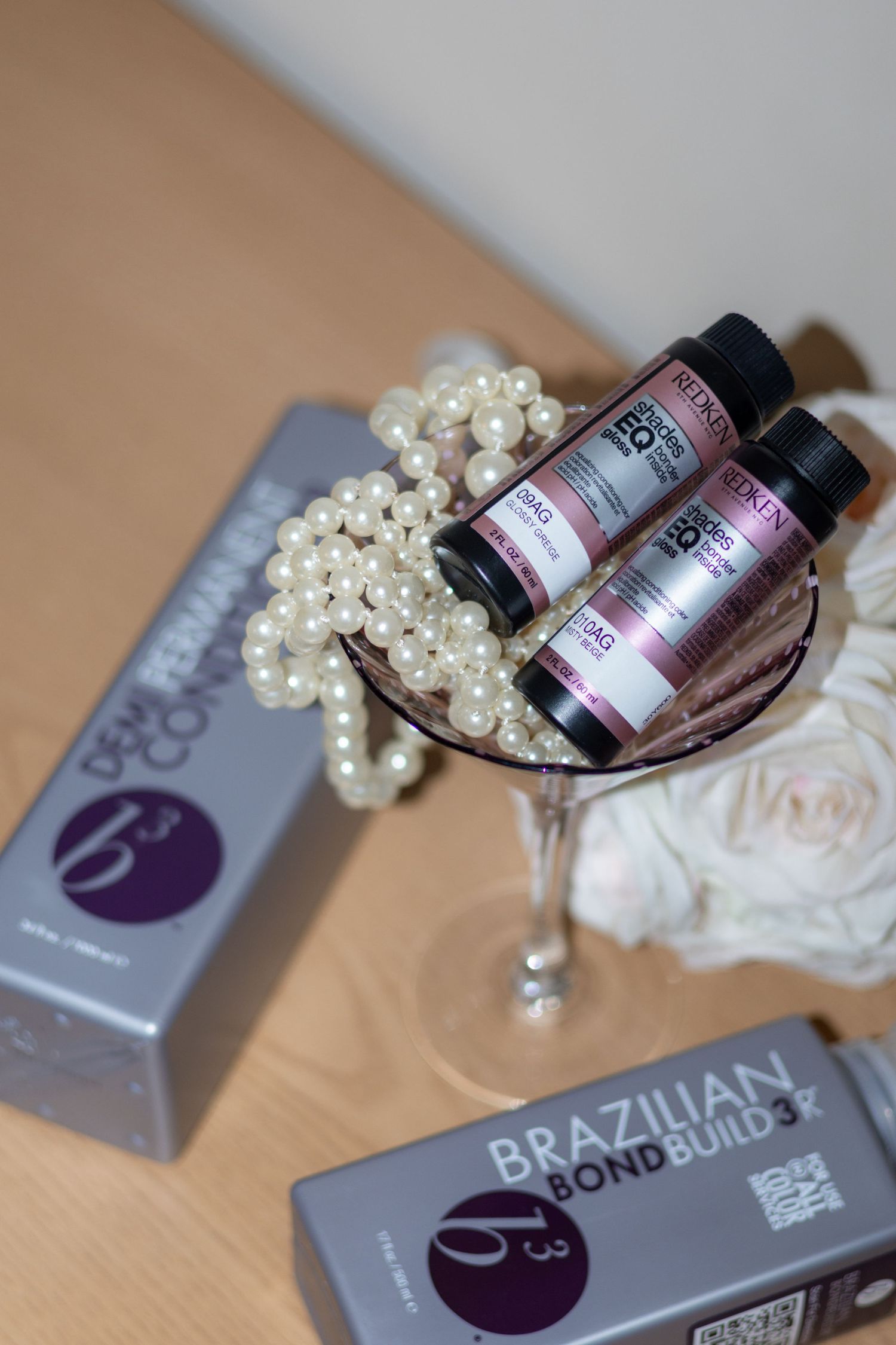 Bottles of Brazilian Bond Builder hair treatment products displayed with pearl necklaces and white fabric on a beige surface.