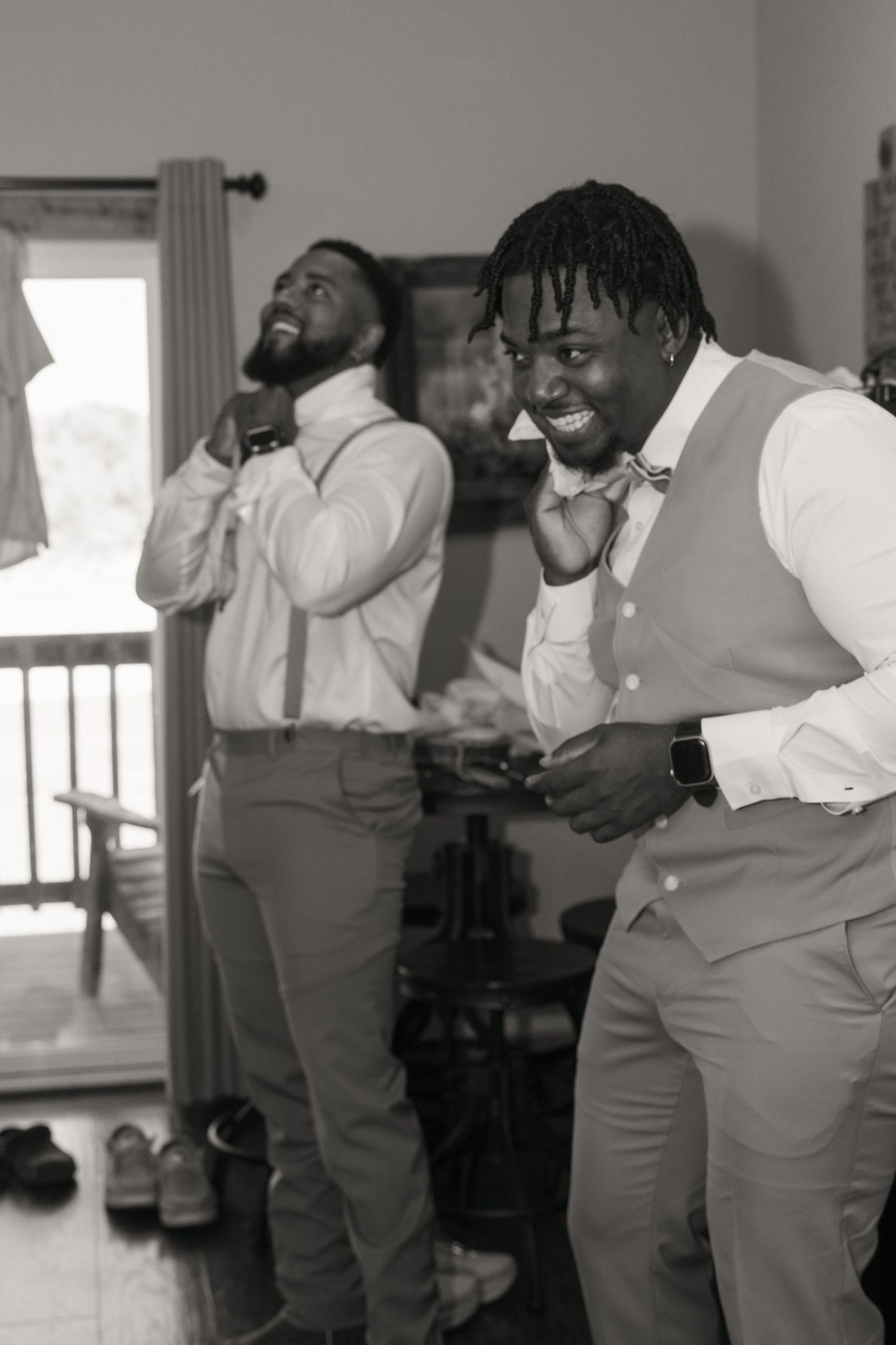 Two groomsmen laughing and adjusting bow ties while getting ready for a wedding ceremony.
