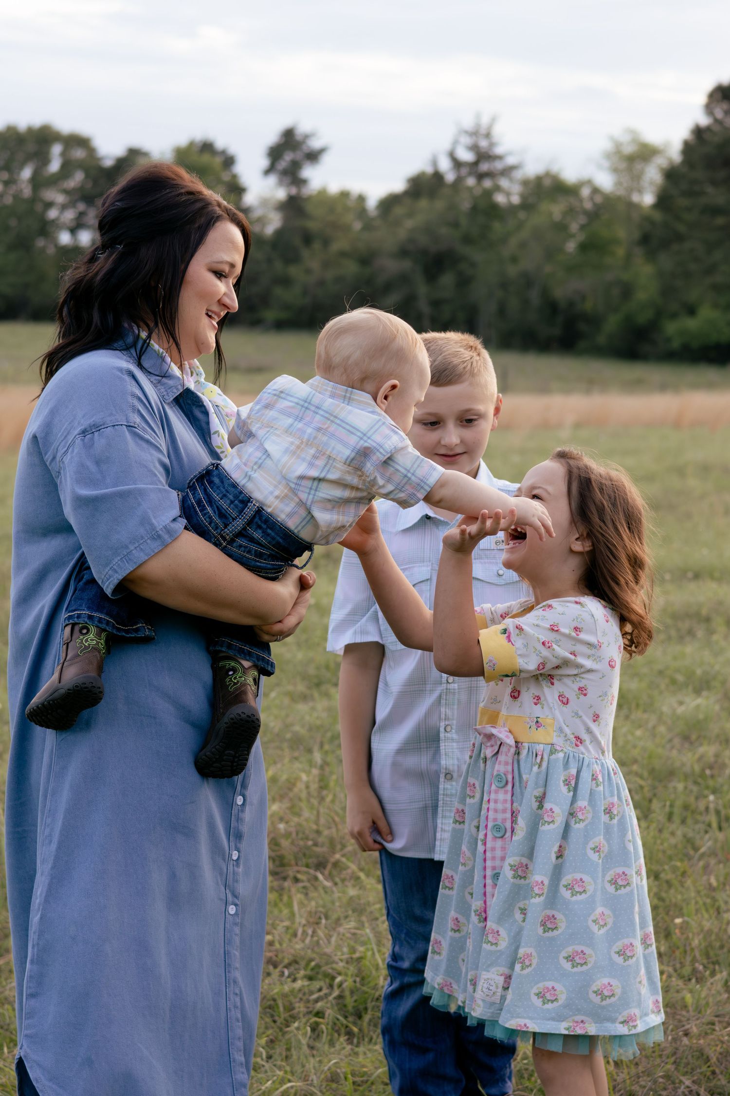 A mother holds her baby while two young children interact playfully in an open field with trees in the background.