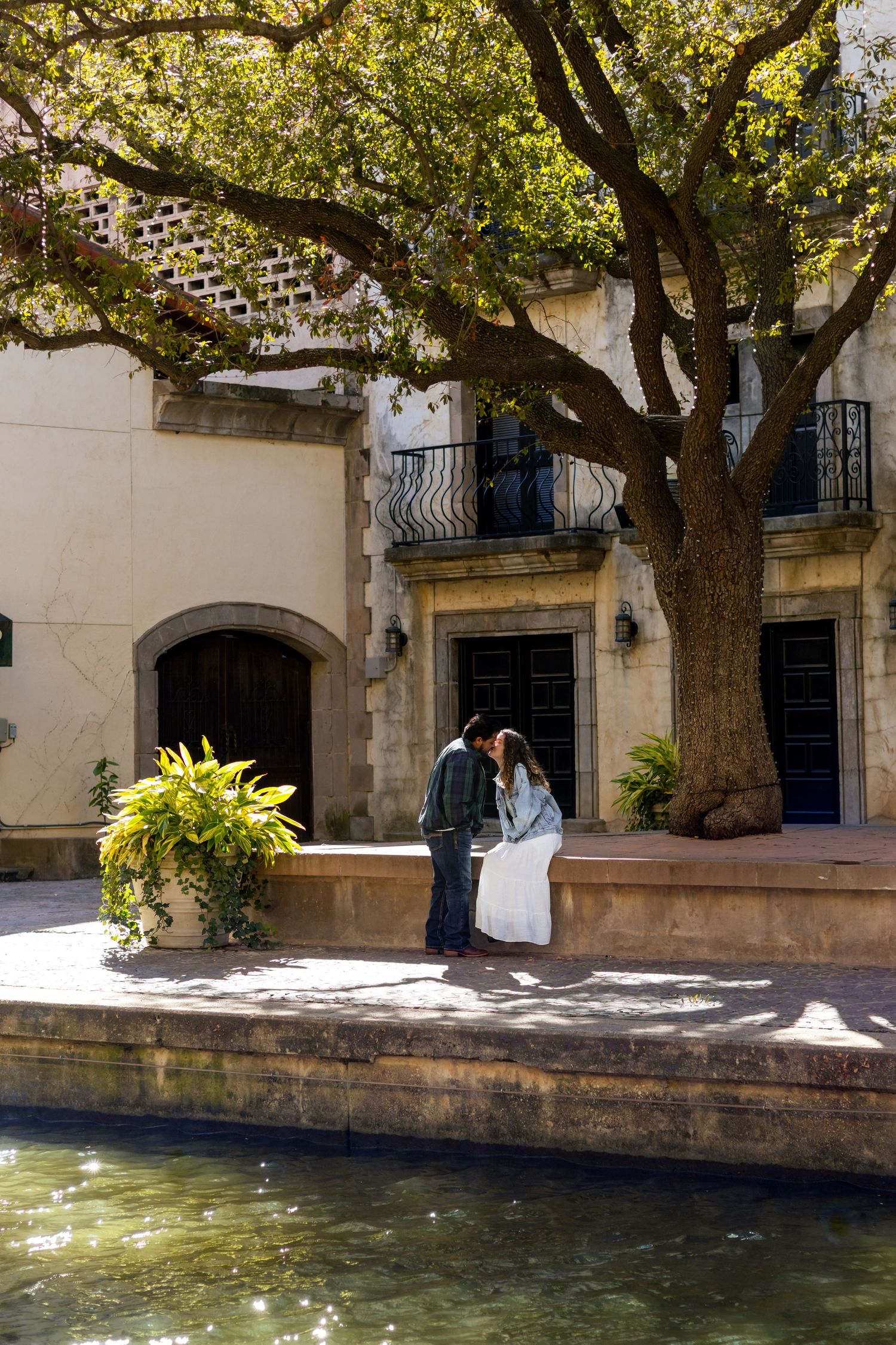 A serene waterfront walkway lined with shady trees and historic stone buildings in a Mediterranean style setting.