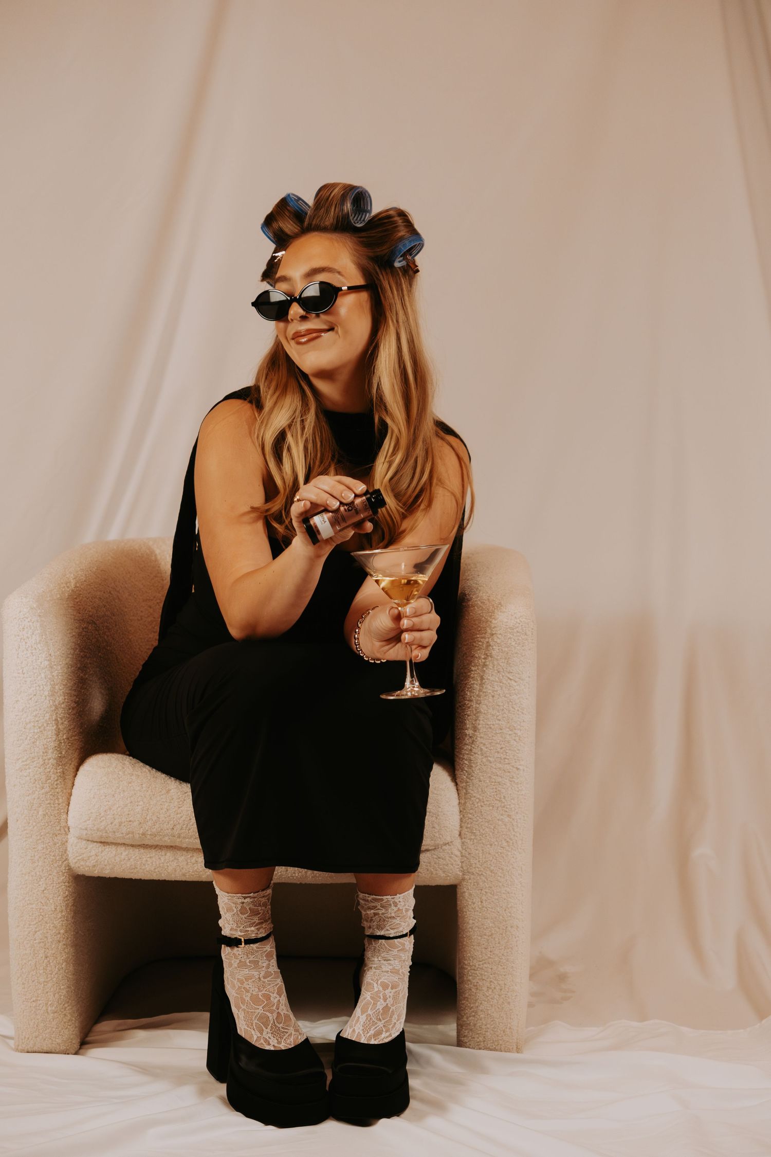 Woman in black dress with hair rollers, sunglasses, socks, and platform shoes sits in chair holding a martini glass.