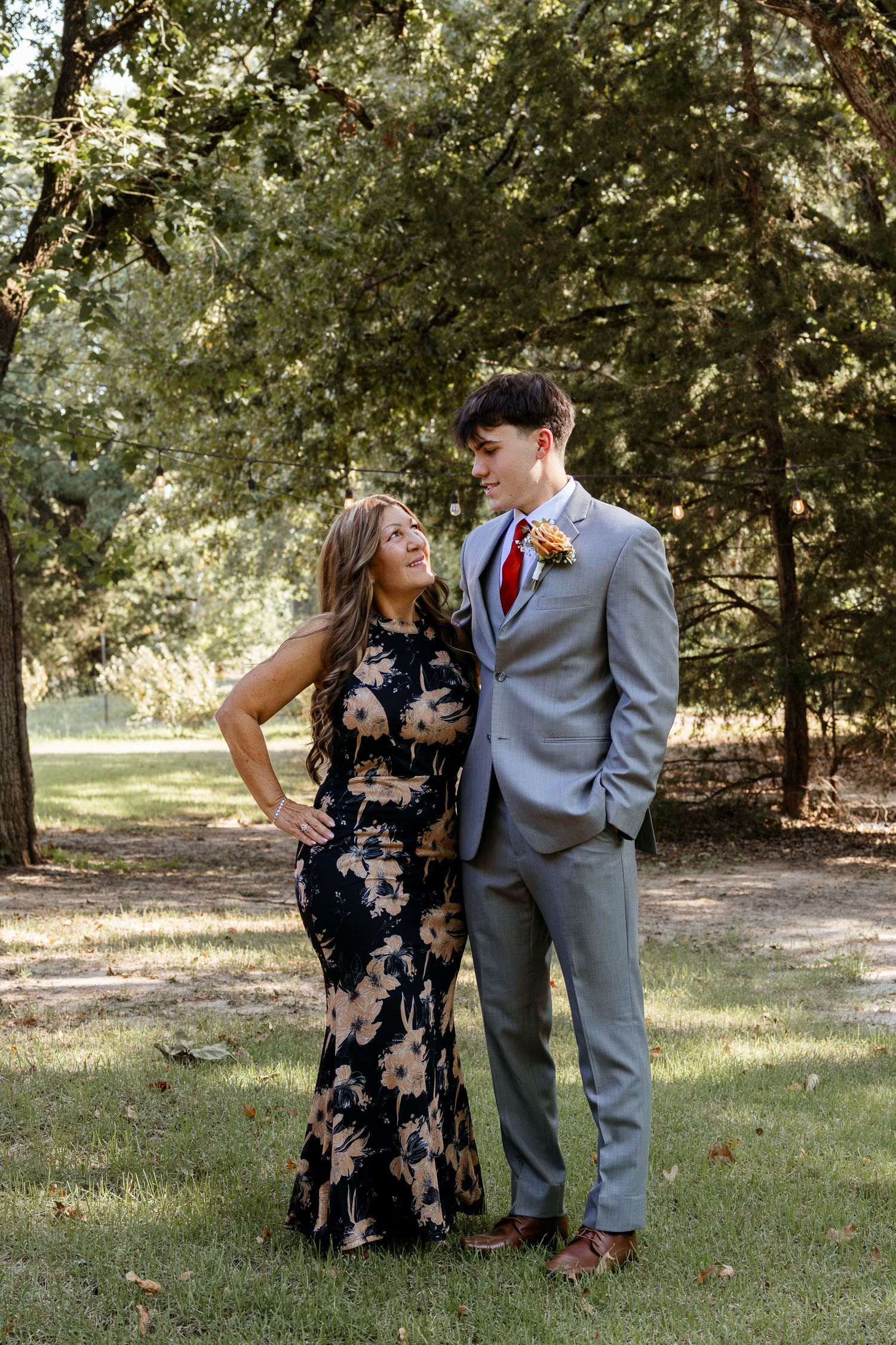 Couple in formal attire pose outdoors under green trees, man in gray suit with boutonniere.