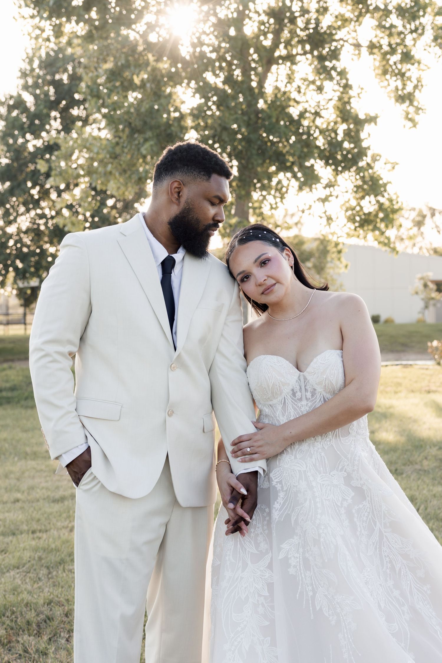 Interracial couple in wedding attire pose outdoors at golden hour, groom in cream suit, bride in strapless lace gown.